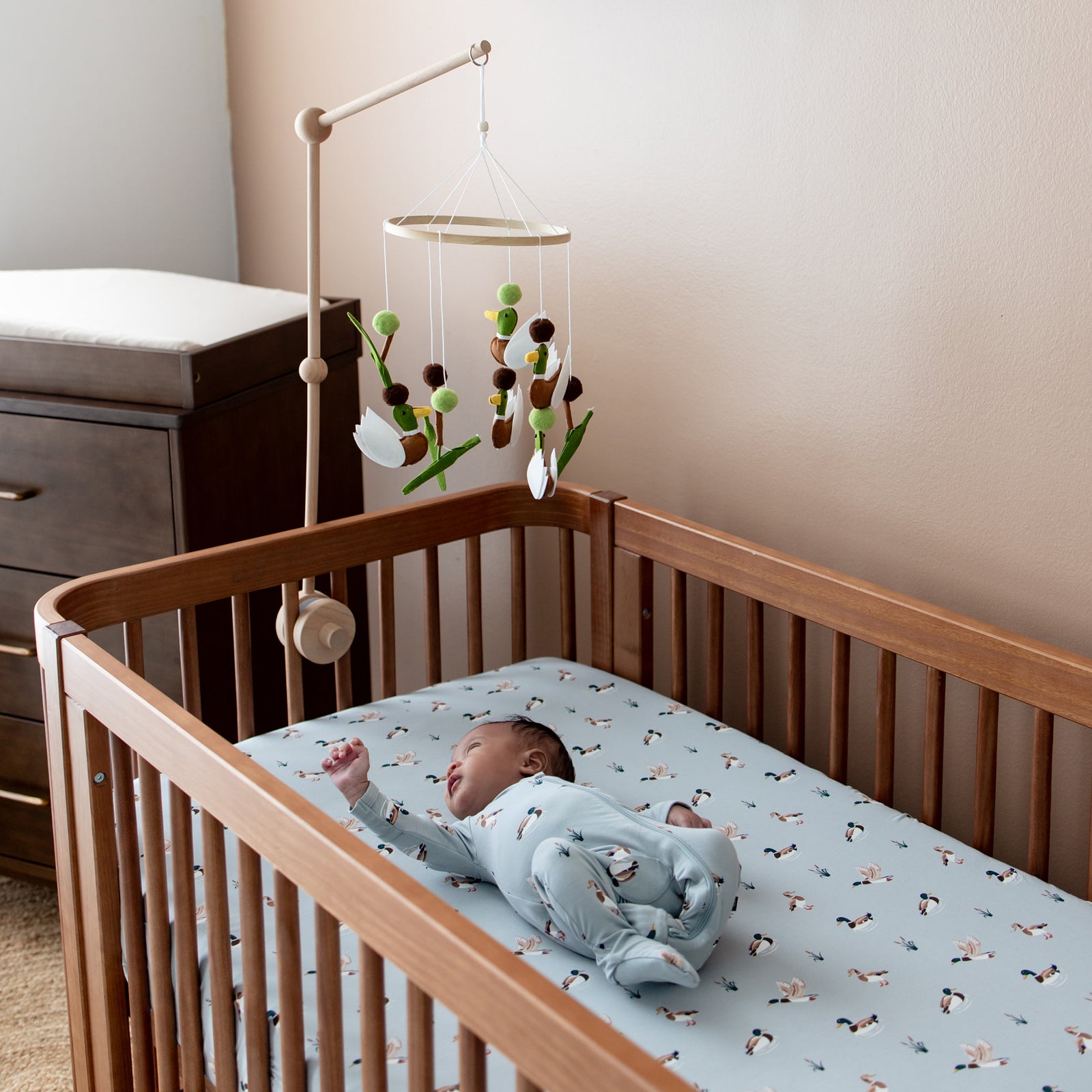 Wooden crib with the Crib Sheet in Mallard Duck on the mattress with an infant laying on the crib sheet wearing a matching zippered footie