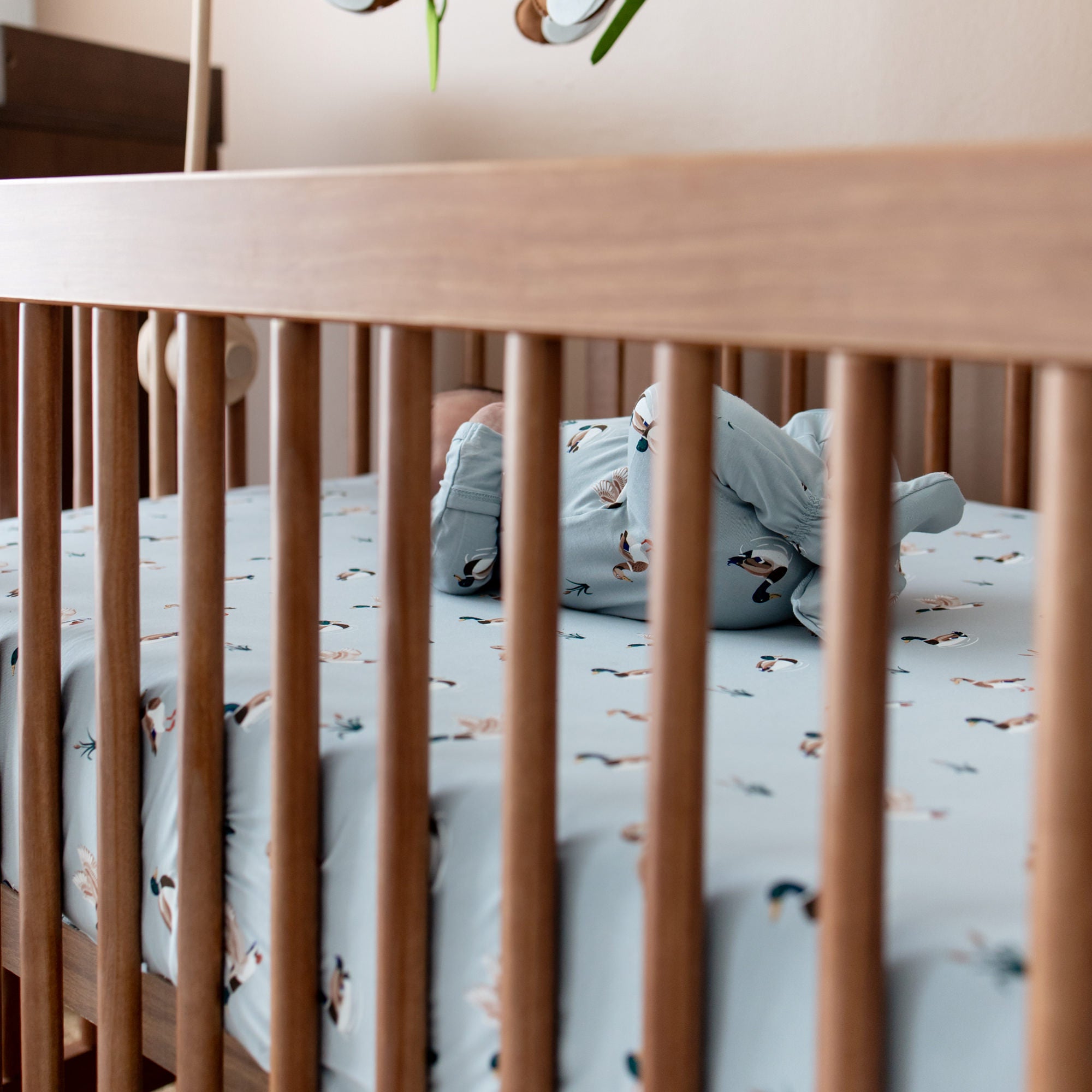 Wide shot of the wooden crib with the Crib Sheet in Mallard Duck on the crib sheet with an infant laying in the crib