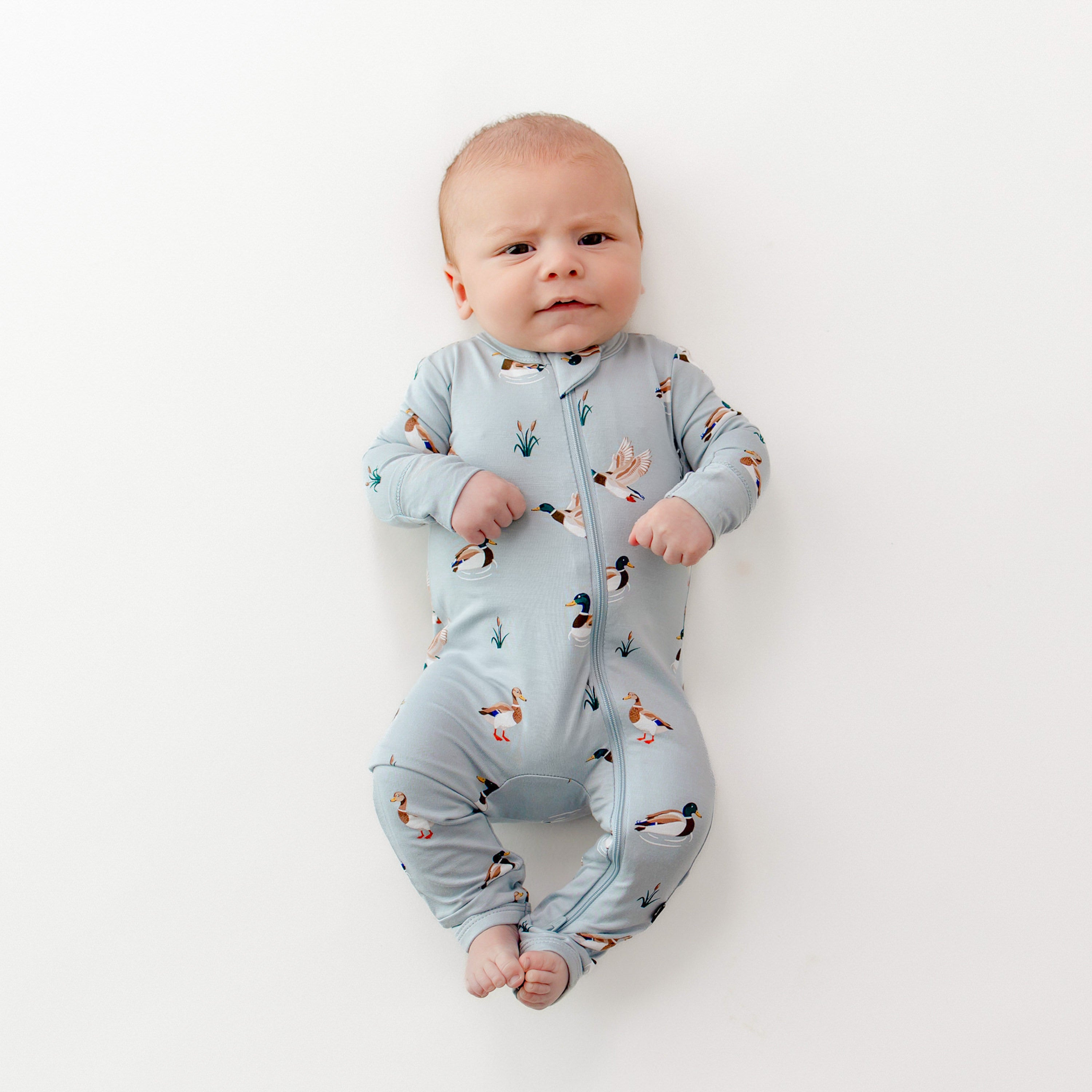 Infant laying on a white surface wearing the Zippered Romper in Mallard Duck