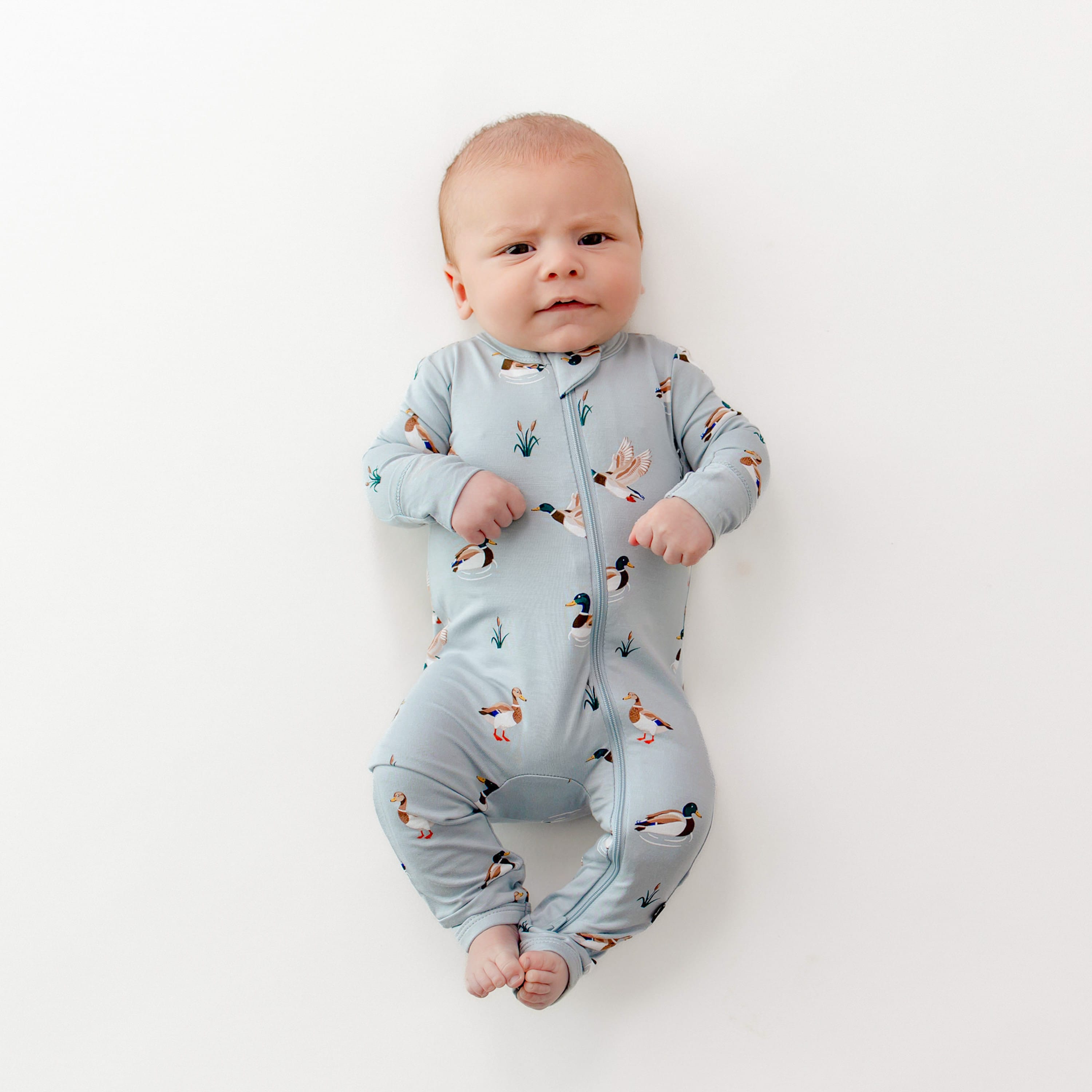 Infant laying on a white surface wearing the Zippered Romper in Mallard Duck