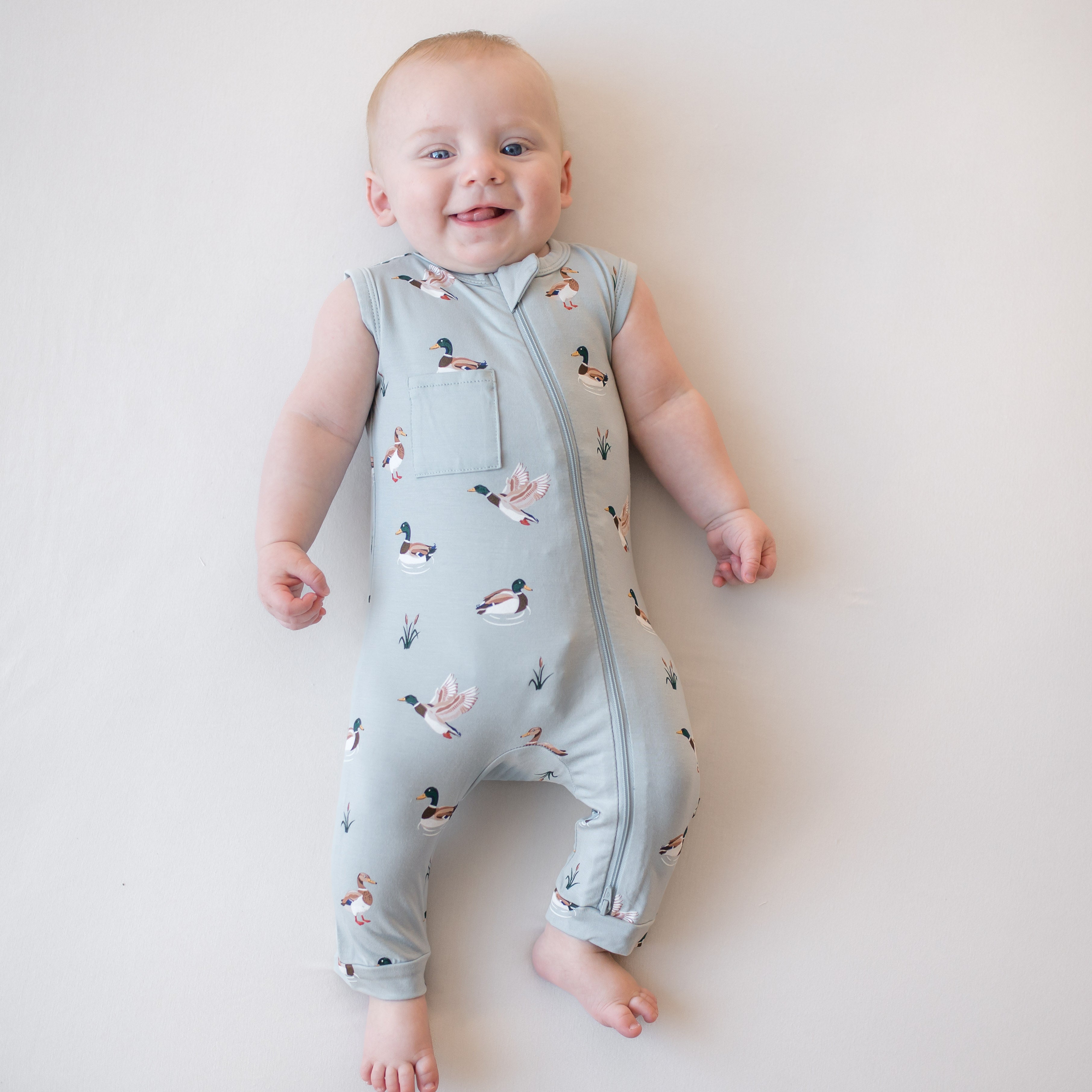Smiling infant laying on a light surface wearing the Zippered Sleeveless Romper in Mallard Duck
