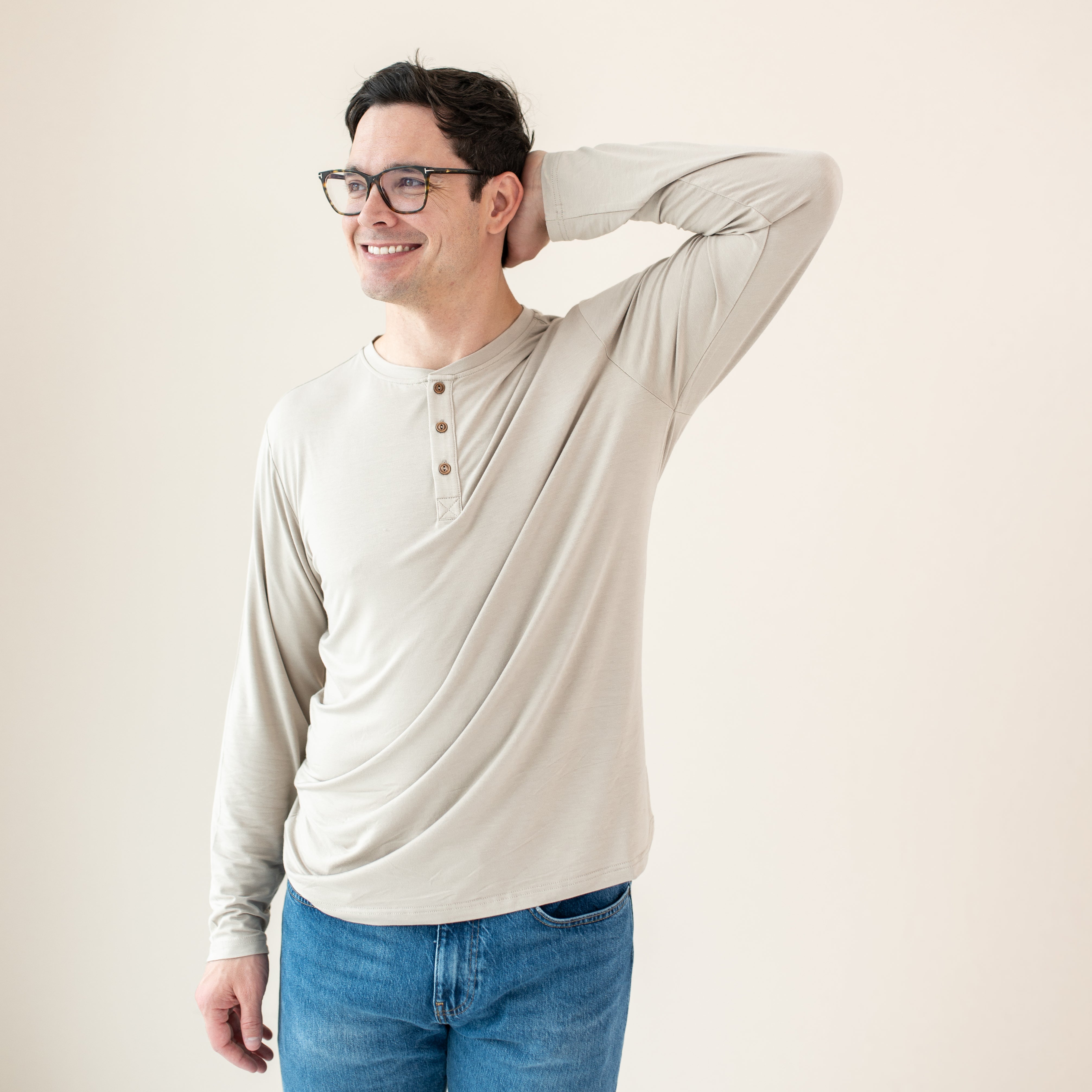 Smiling male model standing with his hand behind is head wearing the Men's Long Sleeve Henley Top in Bisque