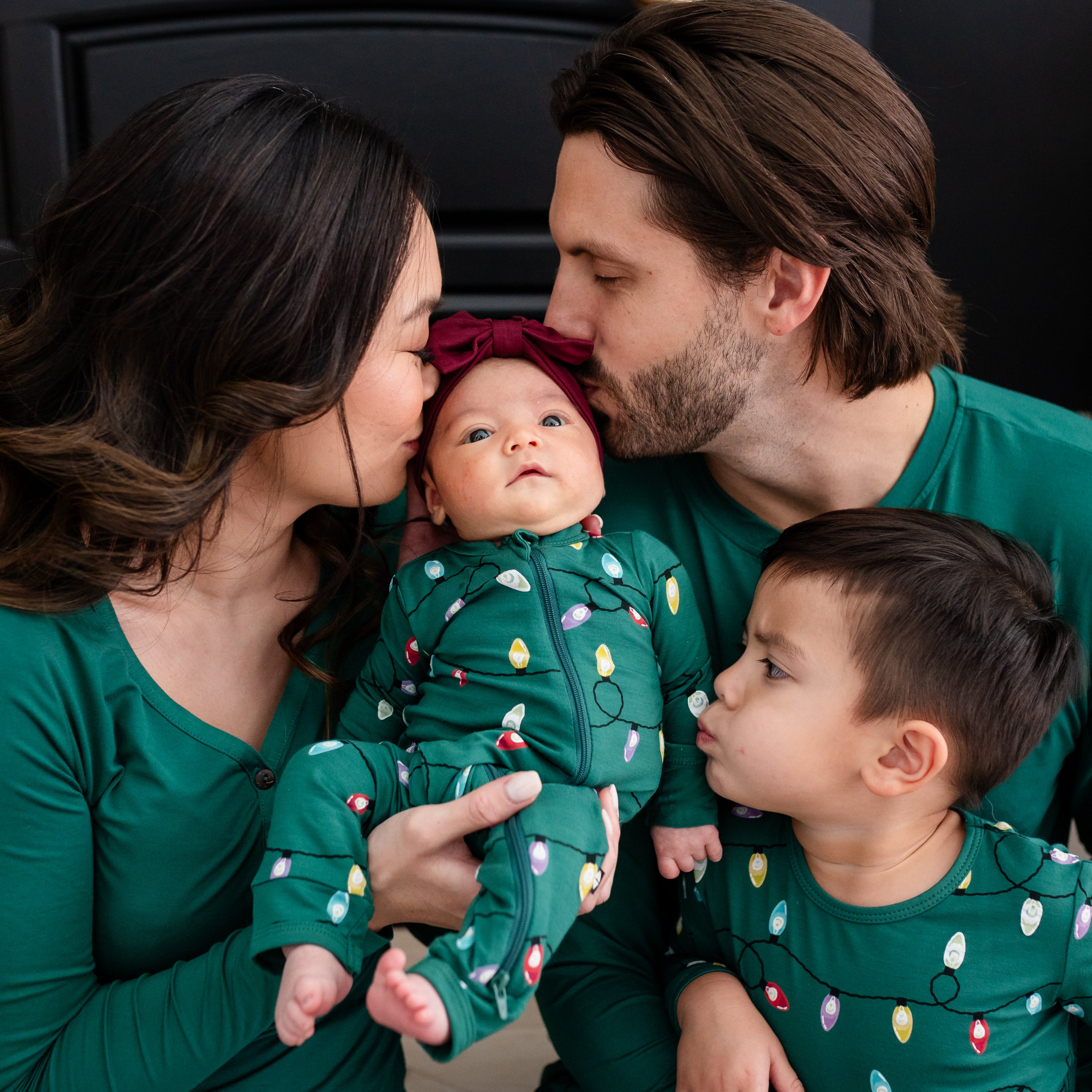 Mother wearing the Emerald women's Henley tee holding newborn baby who is wearing the Zippered Romper in Merry and Bright while mother, father and son all kiss the baby. Father is matching mother wearing an Emerald Henley and oldest child is matching newborn in a long sleeve pajama set