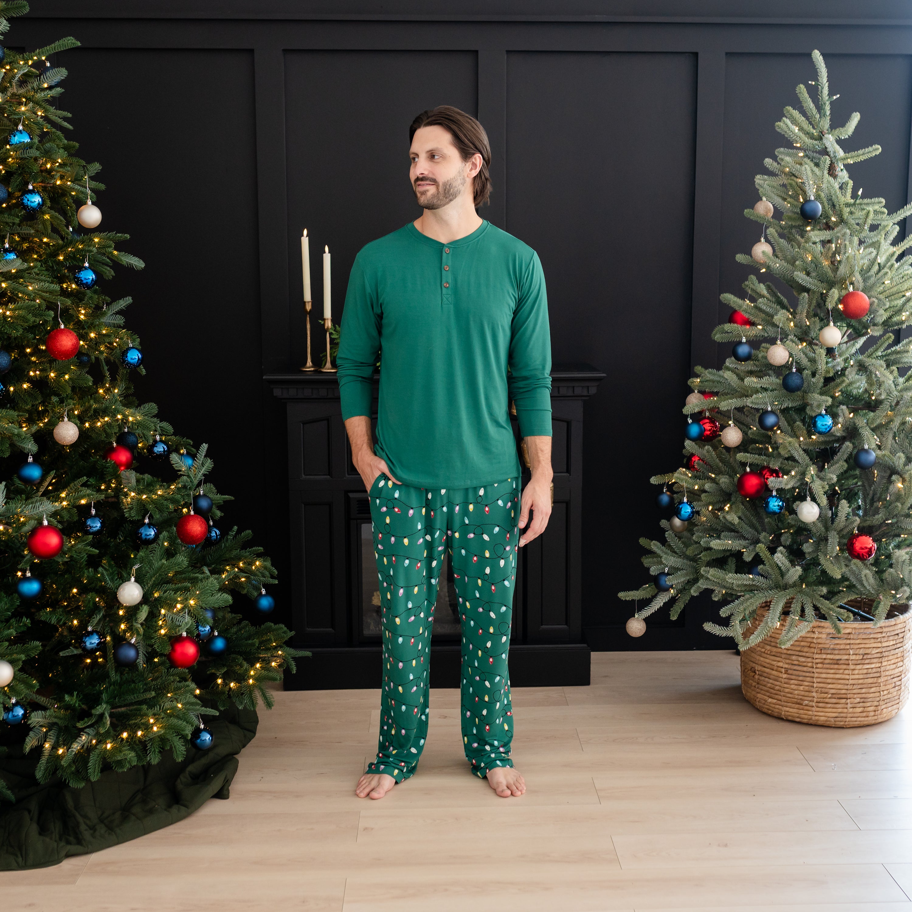 Male model looking off to the side standing with one hand in the pocket of the Men's Lounge Pants in Merry and Bright between two decorated Christmas Trees