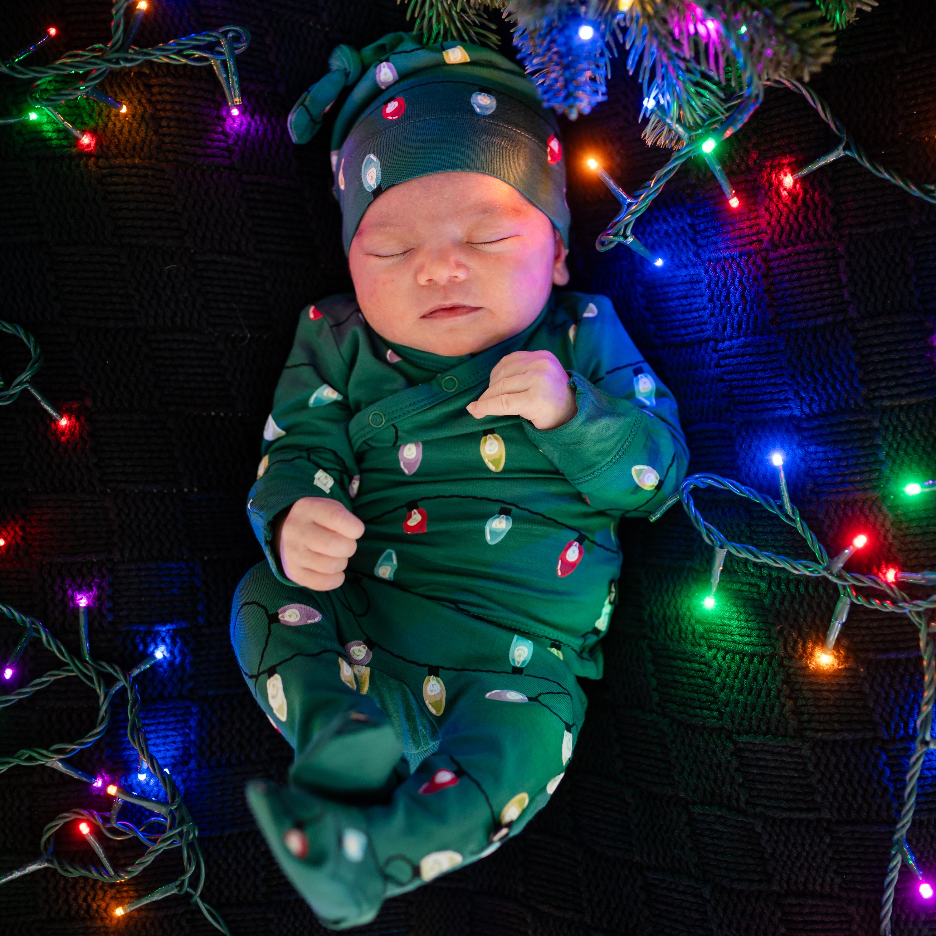 Sleeping newborn sleeping on a Black blanket wearing the Take Me Home Set with Cap in Merry and Bright surround by colored christmas lights