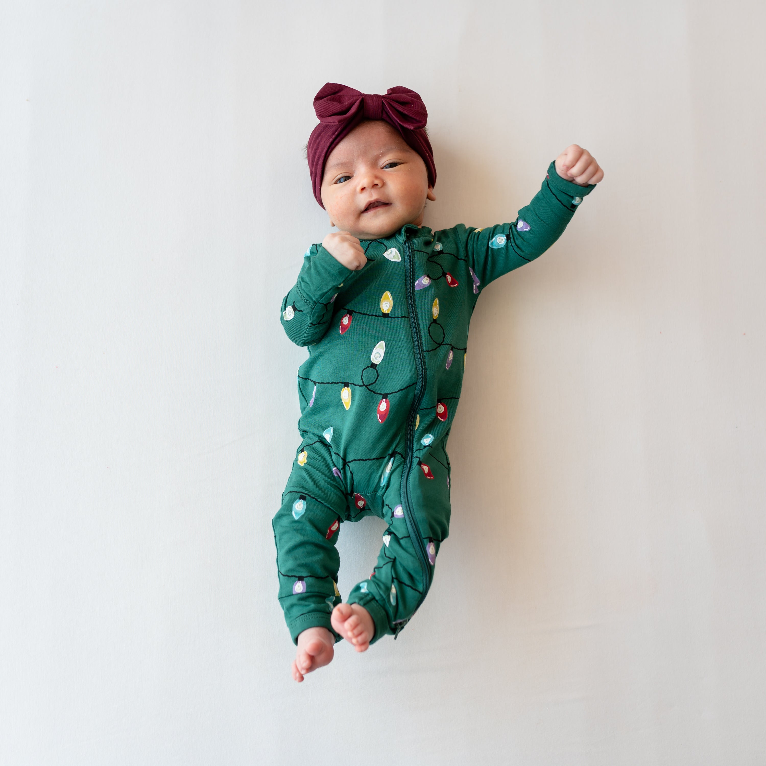 Newborn laying on a cream blanket with her hands in the air wearing the Zippered Romper in Merry and Bright and burgundy bow headband
