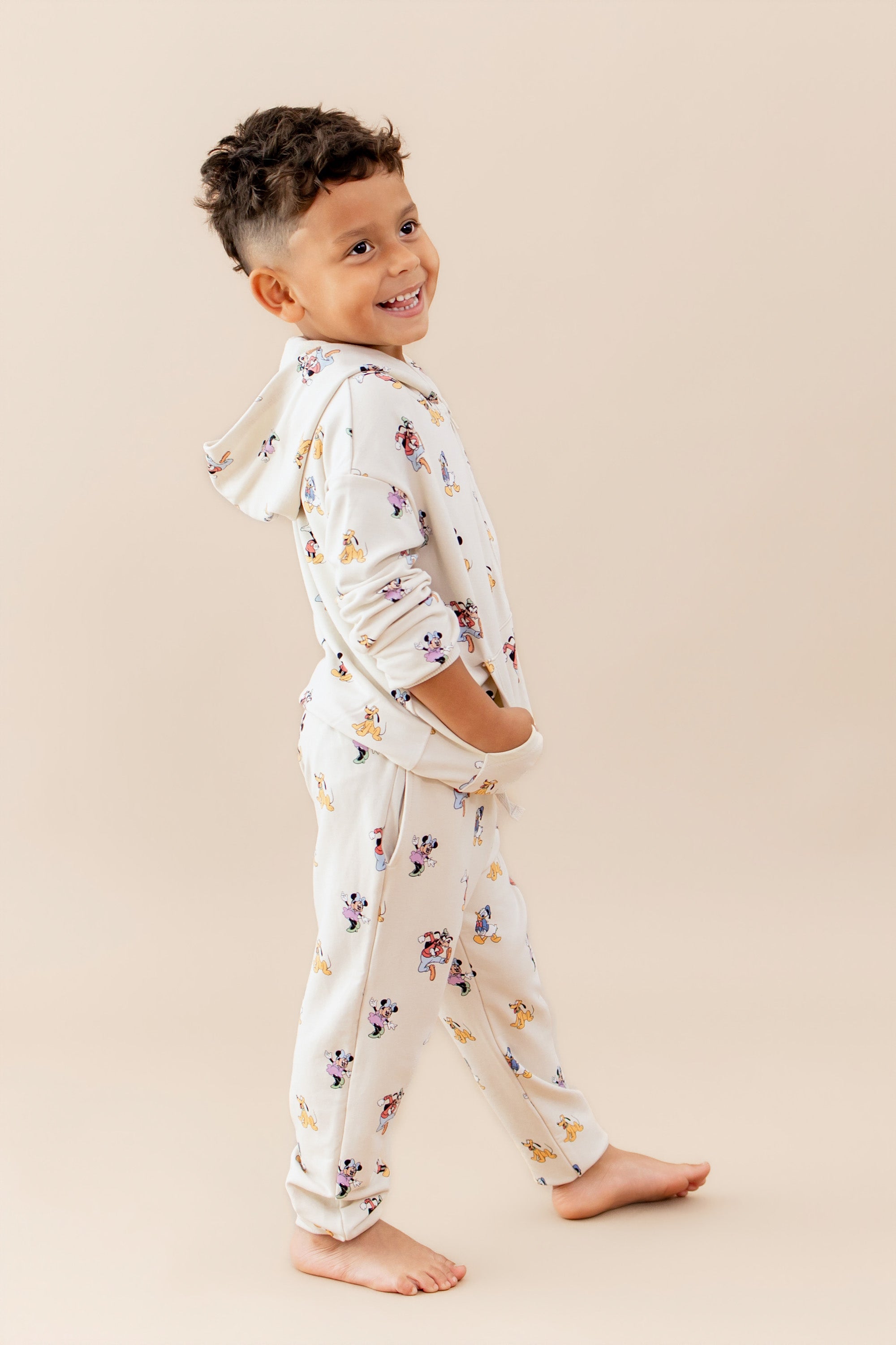 Child walking in a matching sweatsuit with a mickey and friends pattern featuring a zip up hoodie and jogger style pants