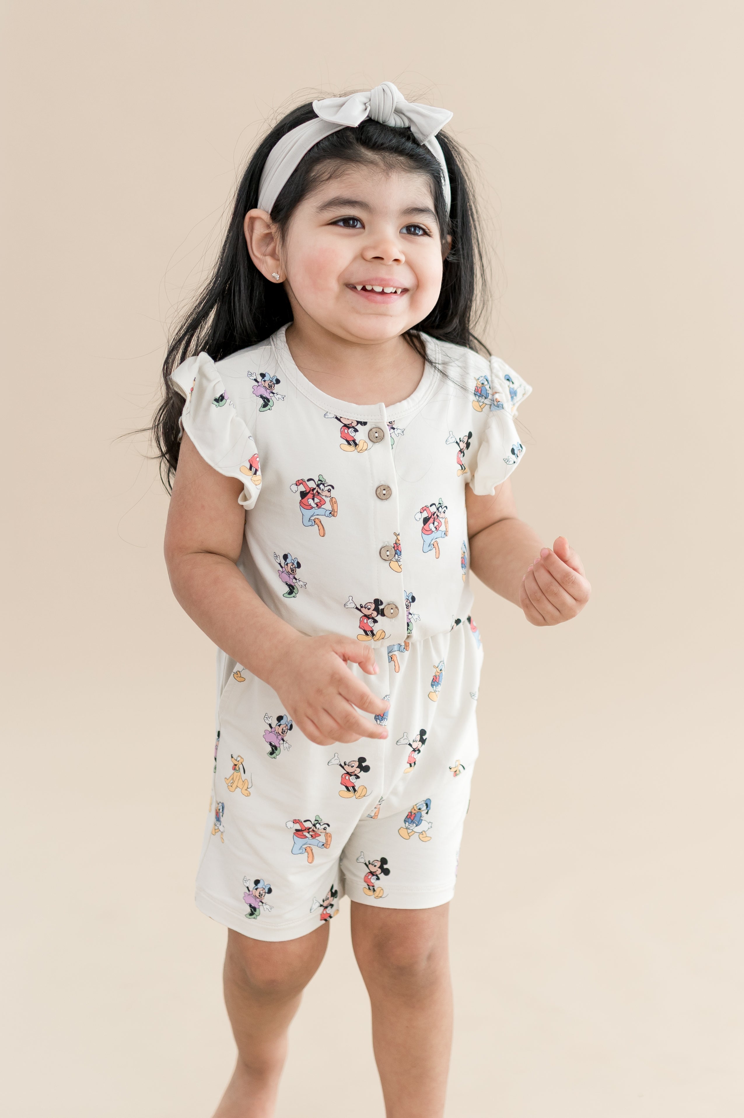 Child wearing mickey mouse and friends printed flutter sleeve romper with buttons on the front and cream bow