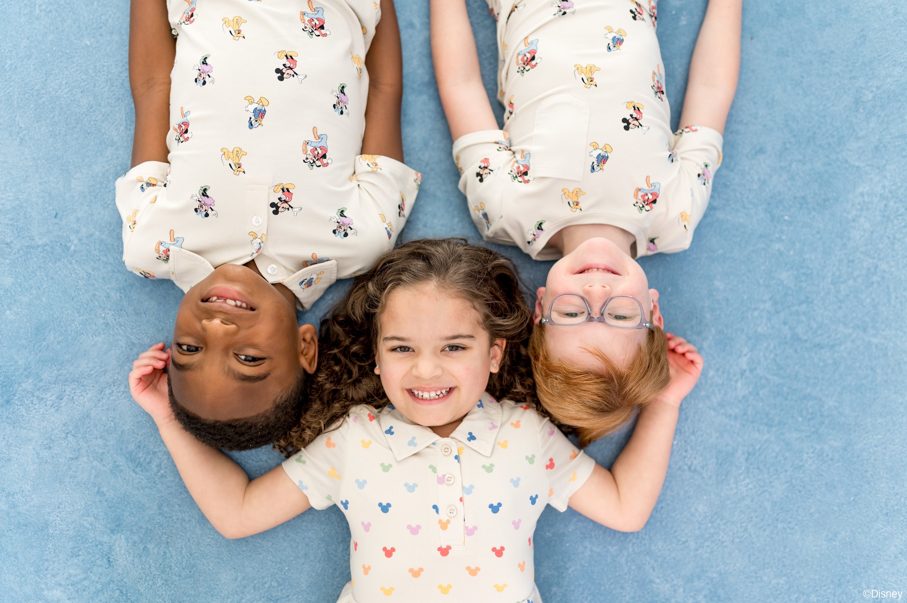 3 children laying on a blue backdrop wearing Kyte's collaboration with Disney featuring mickey and friends and pastel mickey mouse head patterned bamboo rayon apparel