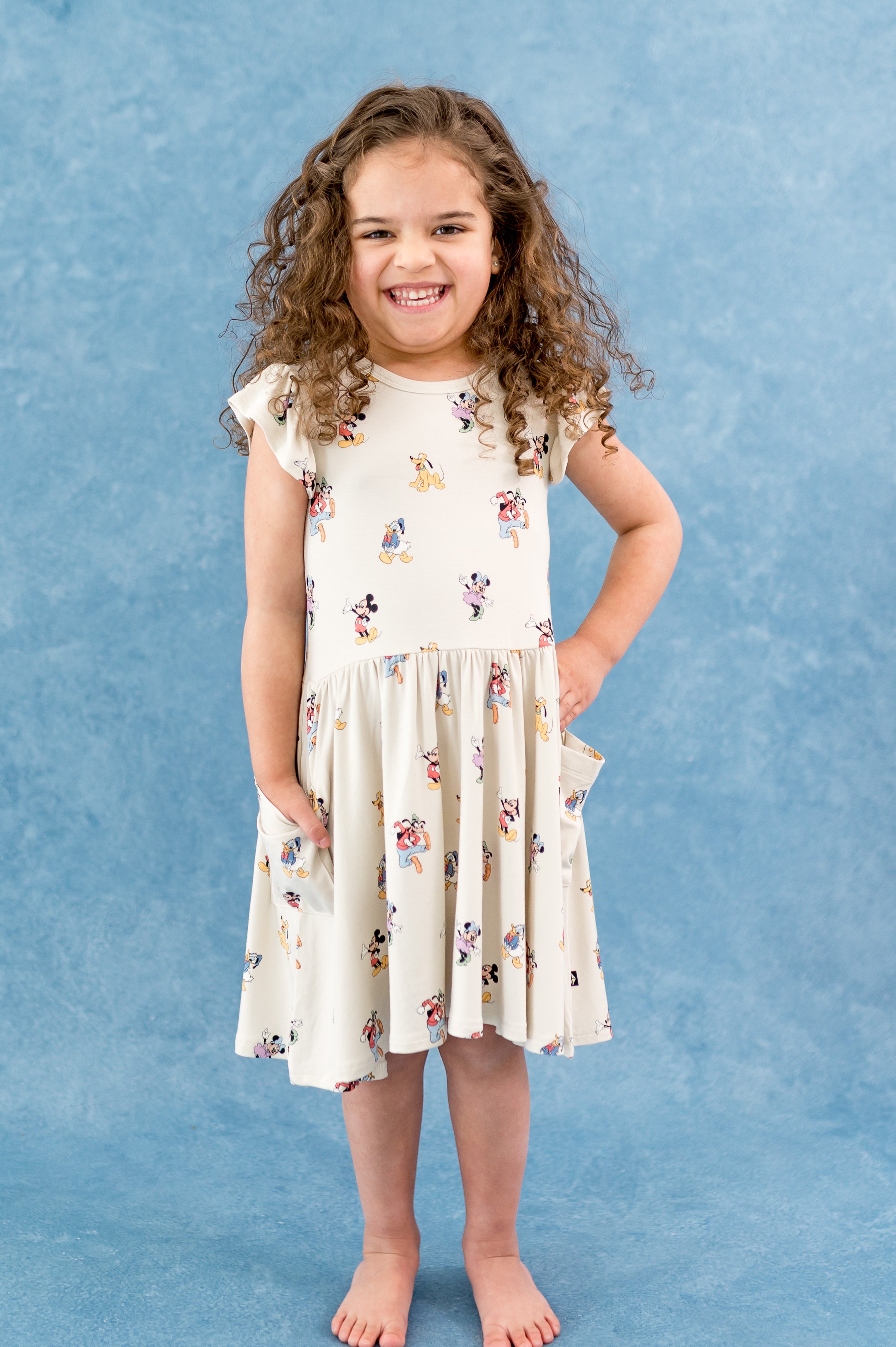 Girl wearing dress with flutter sleeve and pockets in a mickey and friends character pattern with a blue background
