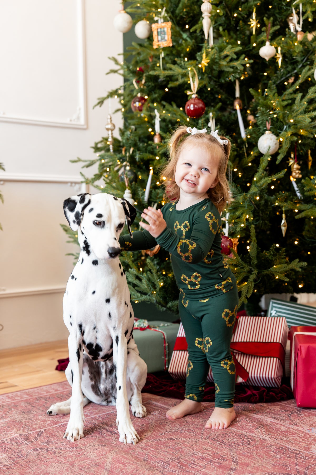 A young child wearing a green Christmas-themed pajama set with gold mickey symbols stands next to a Dalmatian dog in front of a decorated Christmas tree, with wrapped gifts underneath.