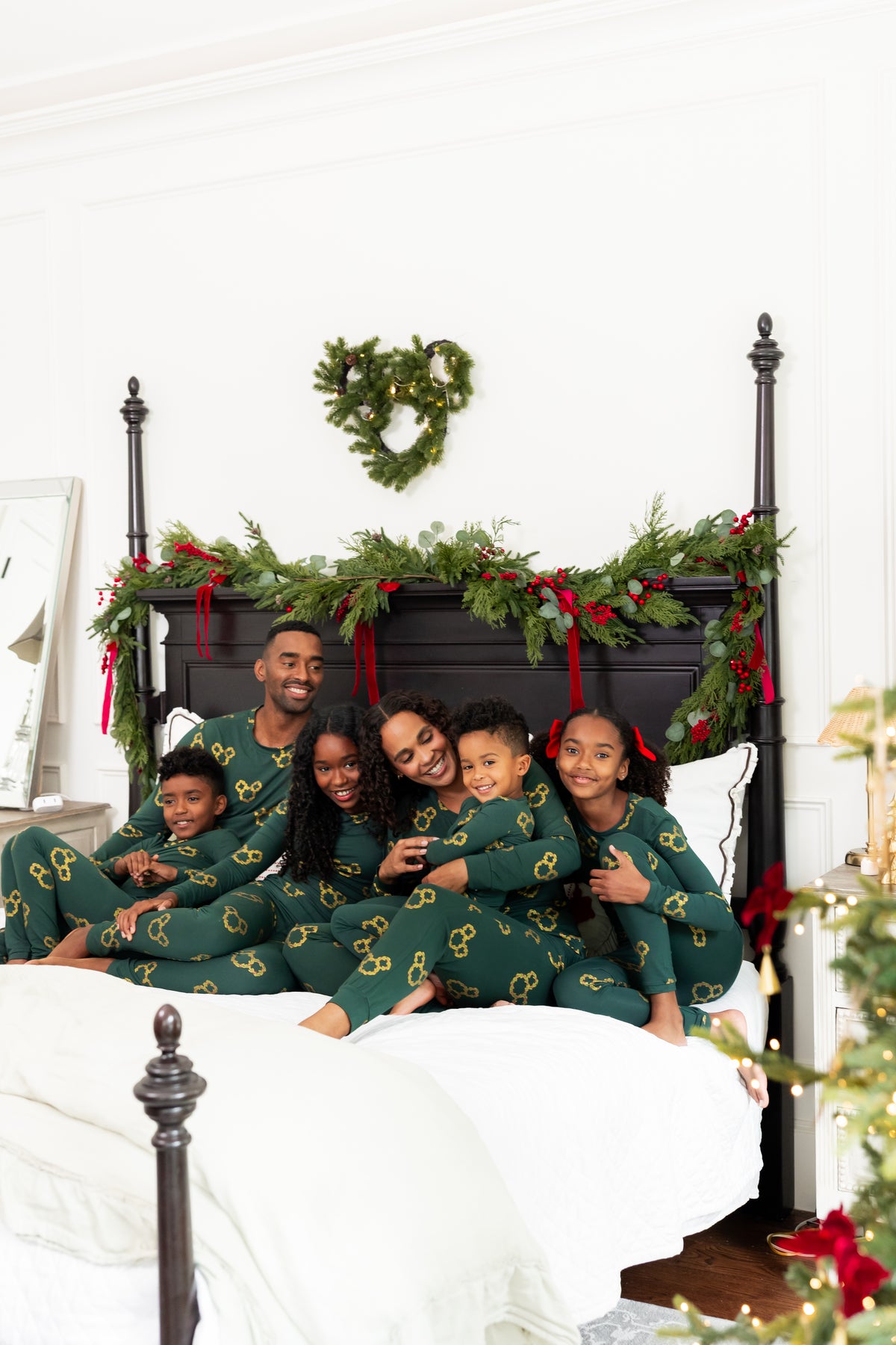 A family of six wearing matching dark green Christmas-themed mickey wreath pajamas with gold accents, sitting together on a bed with a black metal headboard decorated with a green pine garland and red ribbons.