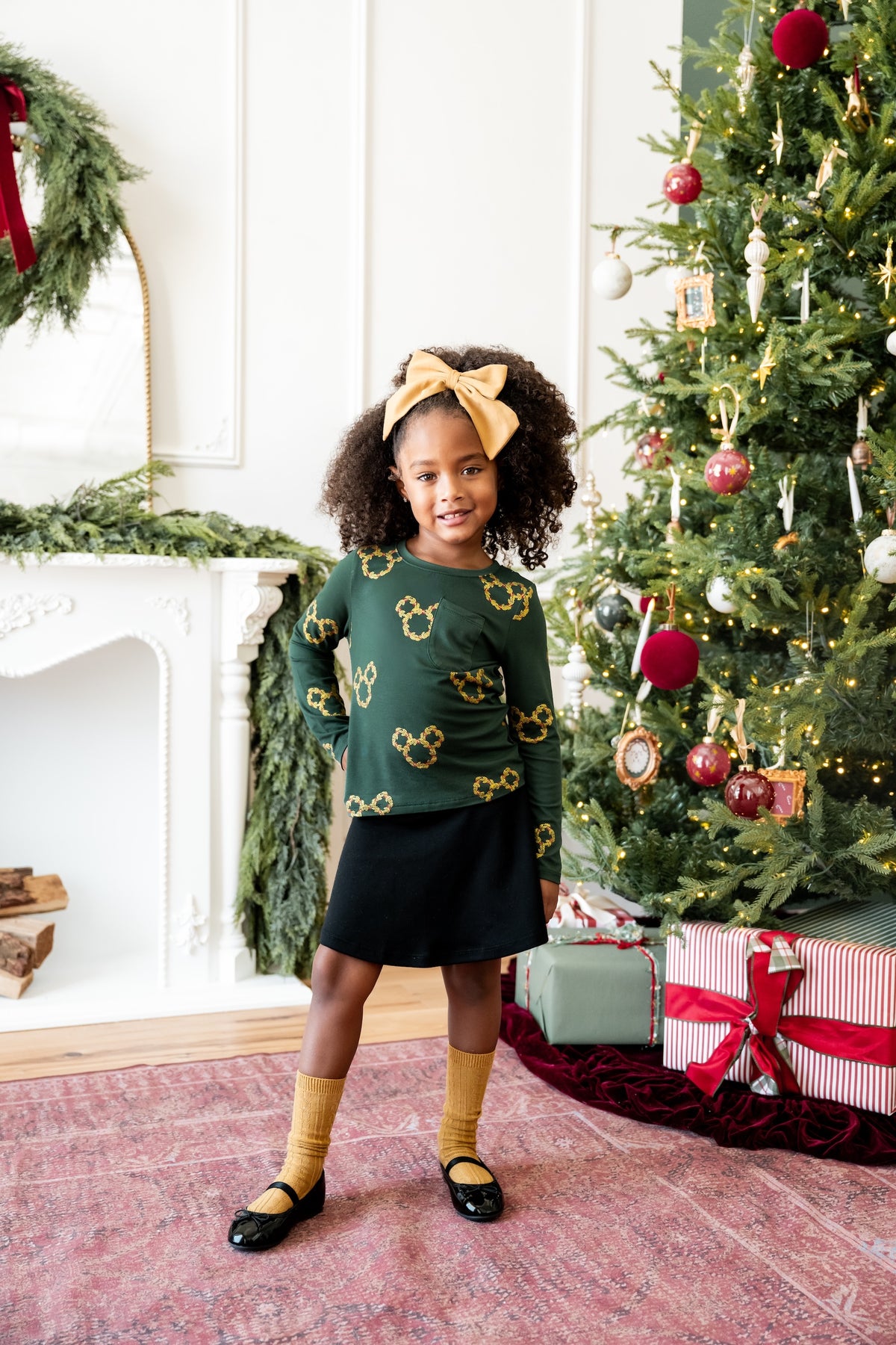 Girl wearing Long Sleeve Toddler Crew Neck Tee in Mickey Christmas Wreath print over black skirt standing beside the Christmas tree