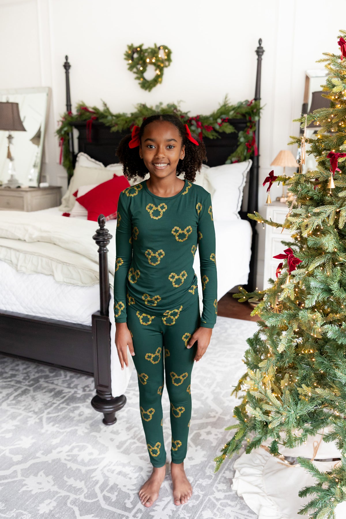 A young girl smiling in a green long-sleeve pajama set with gold Mickey Mouse head patterns, standing in a decorated bedroom next to a Christmas tree.