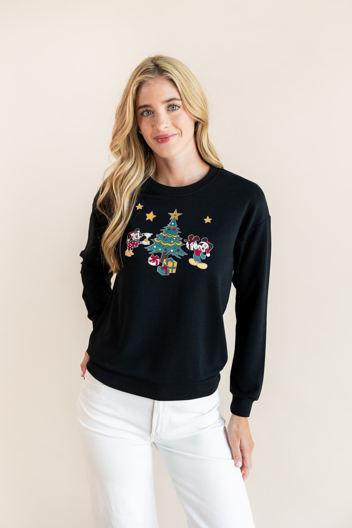 Woman wearing Black Women's Sweatshirt in Mickey & Friends Christmas print in the center over white pants on a beige background