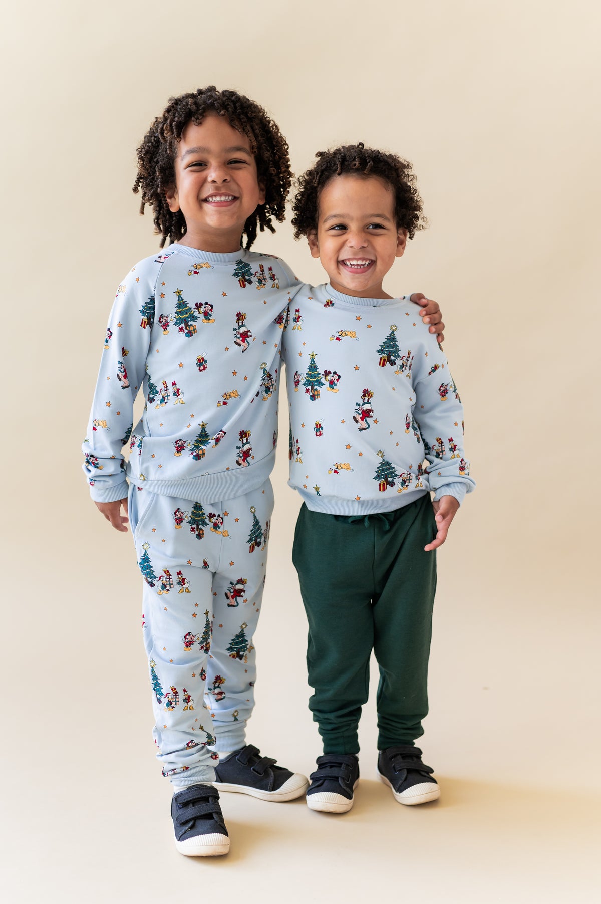 Two young children standing side by side wearing matching light blue Christmas-themed long-sleeve loungewear with a festive mickey print, against a solid beige background.