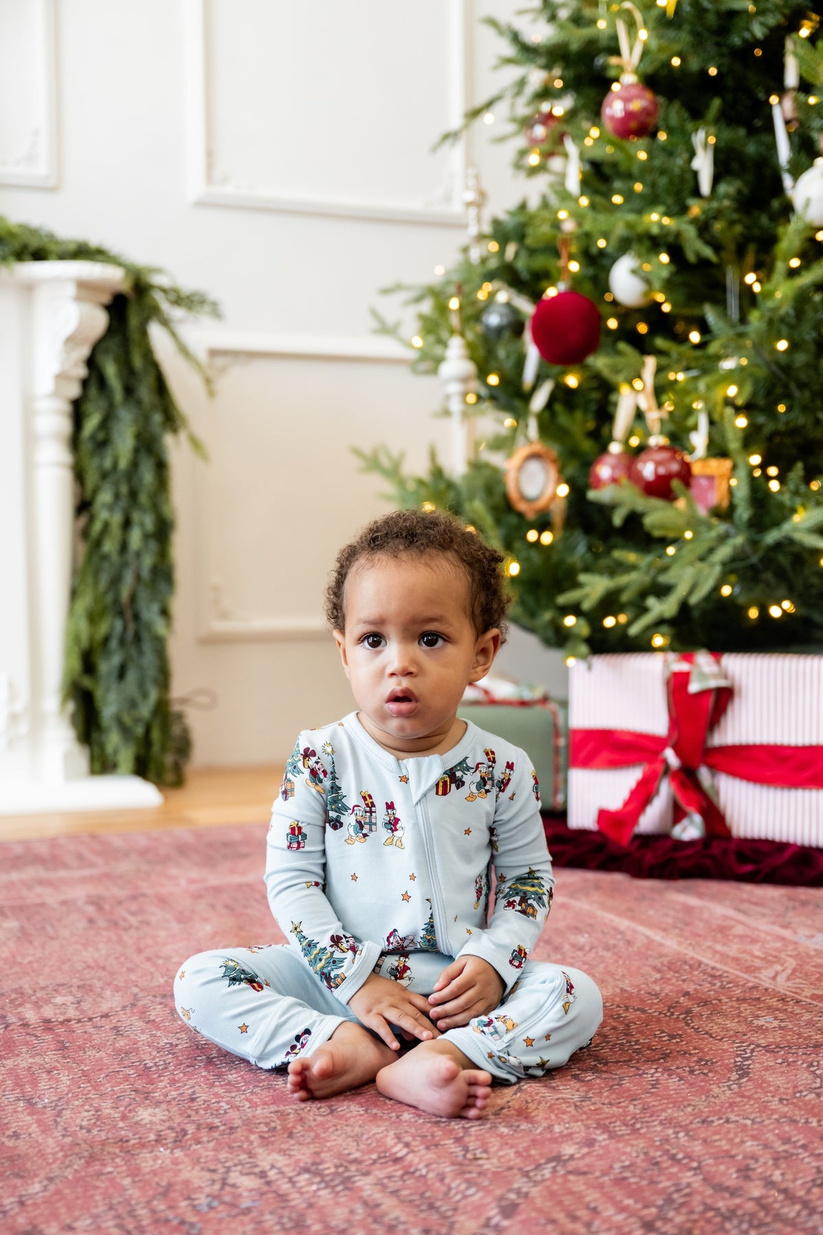 Boy sitting in front of Christmas Tree and presents wearing Zippered Romper in Mickey & Friends Christmas print