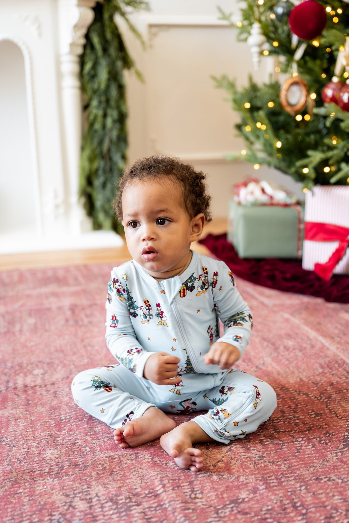 Boy sitting in front of Christmas Tree and presents wearing Zippered Romper in Mickey & Friends Christmas print