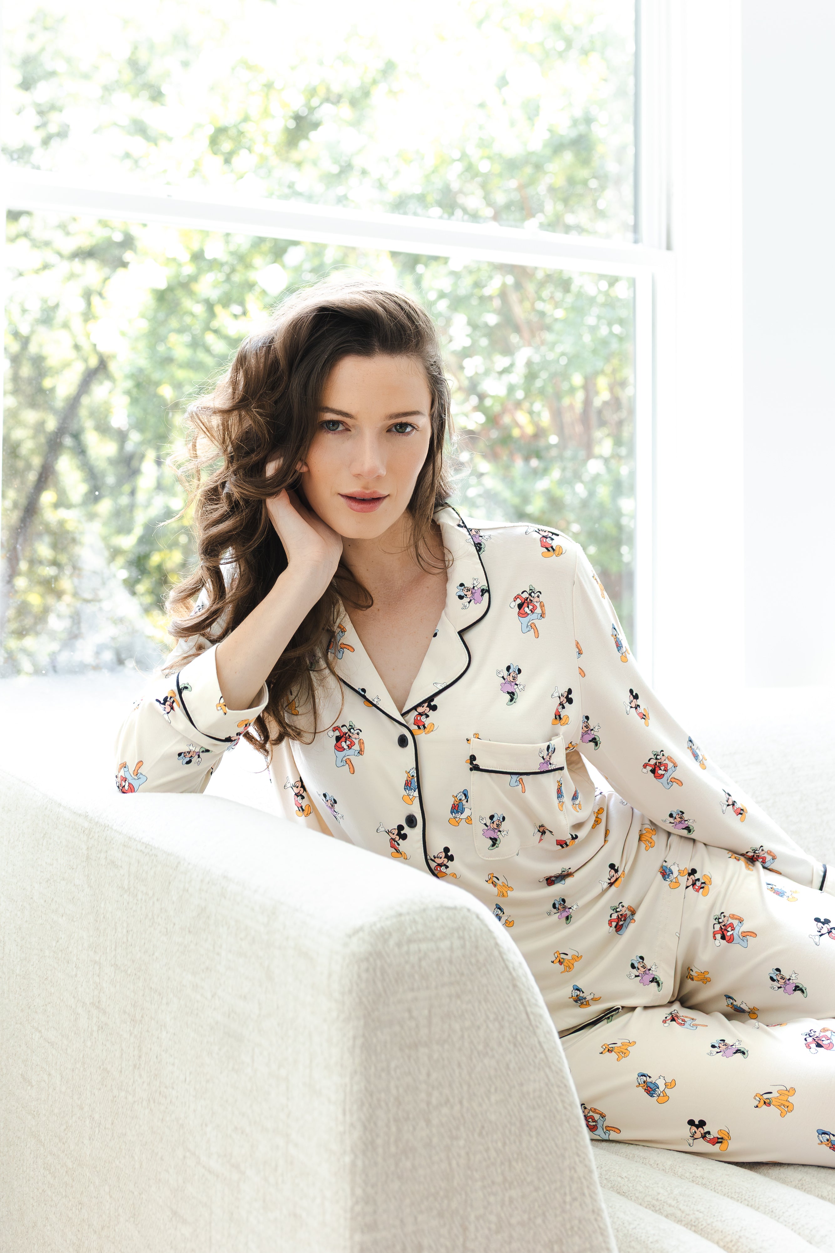 Model sitting on white sofa wearing Long-Sleeved Women's Pajama Set in Mickey Mouse & Friends print