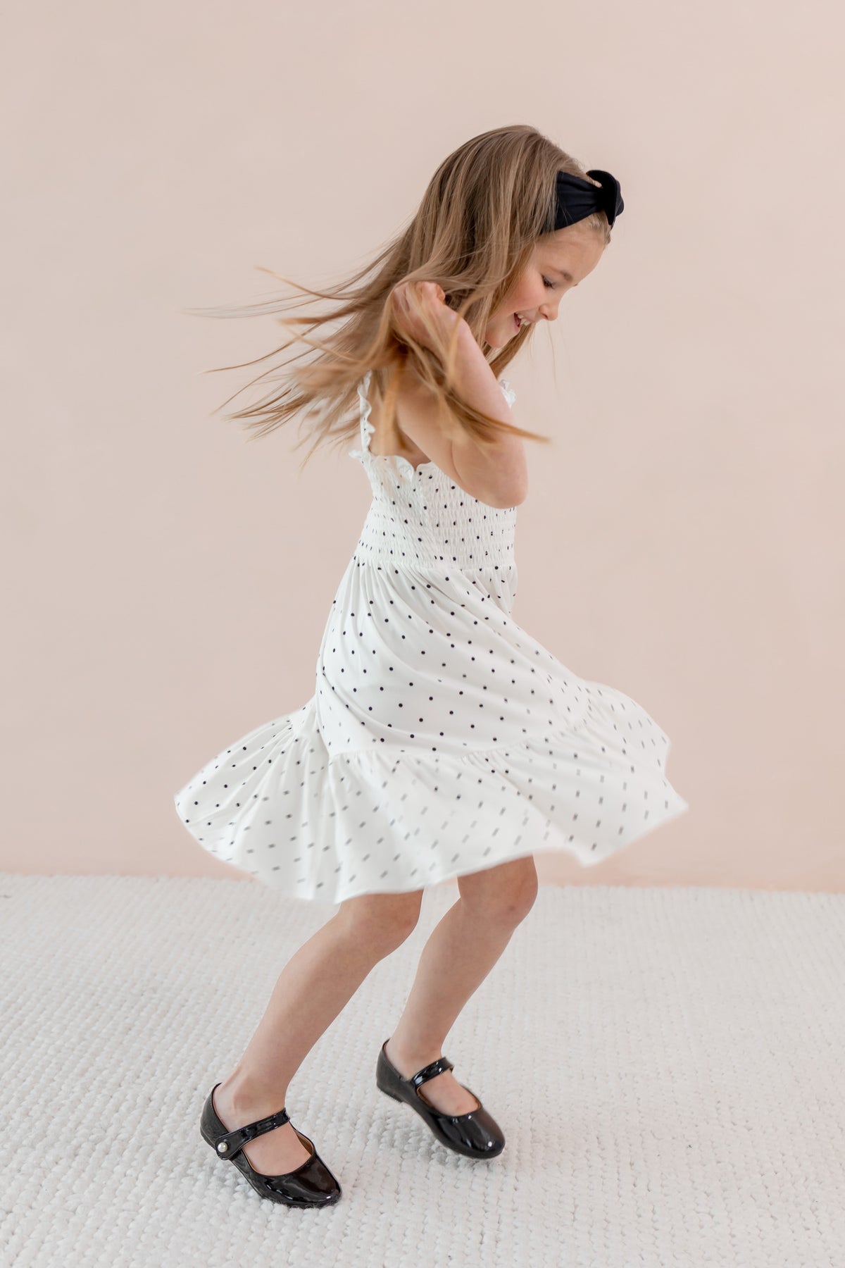 Child in Smocked Dress with Midnight Polka Dot print wearing Black bow headband and shoes on a beige background