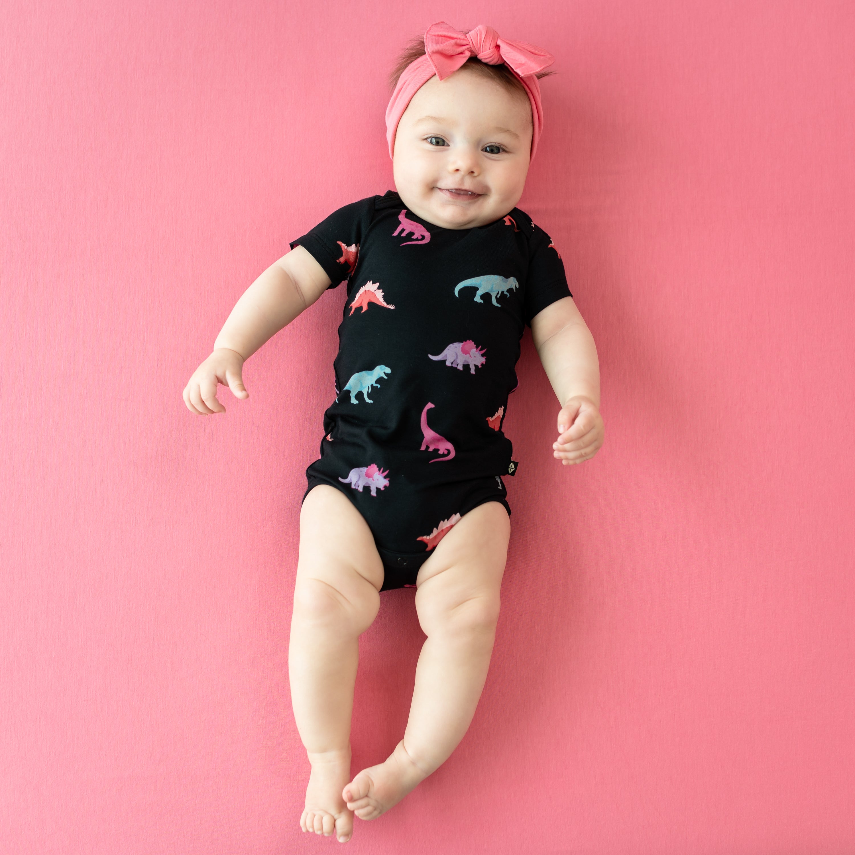 Smiling infant modeling Midnight Roar Short Sleeve Bodysuit wearing a Guava Bow