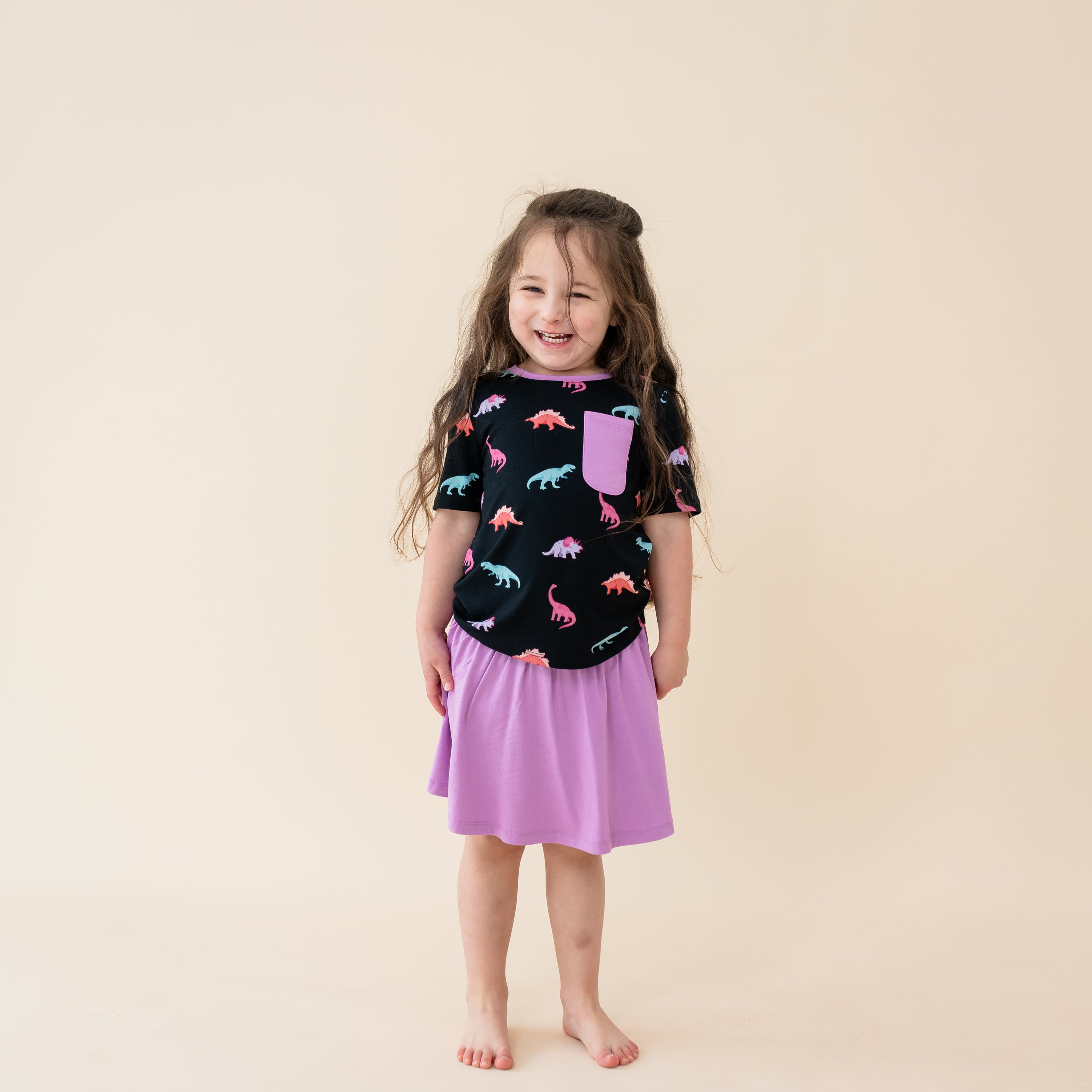 Smiling girl in Midnight Roar Sort Sleeve Toddler Crew Neck Tee. The shirt has a pocket accent in the color Poi and paired with a Poi Skort