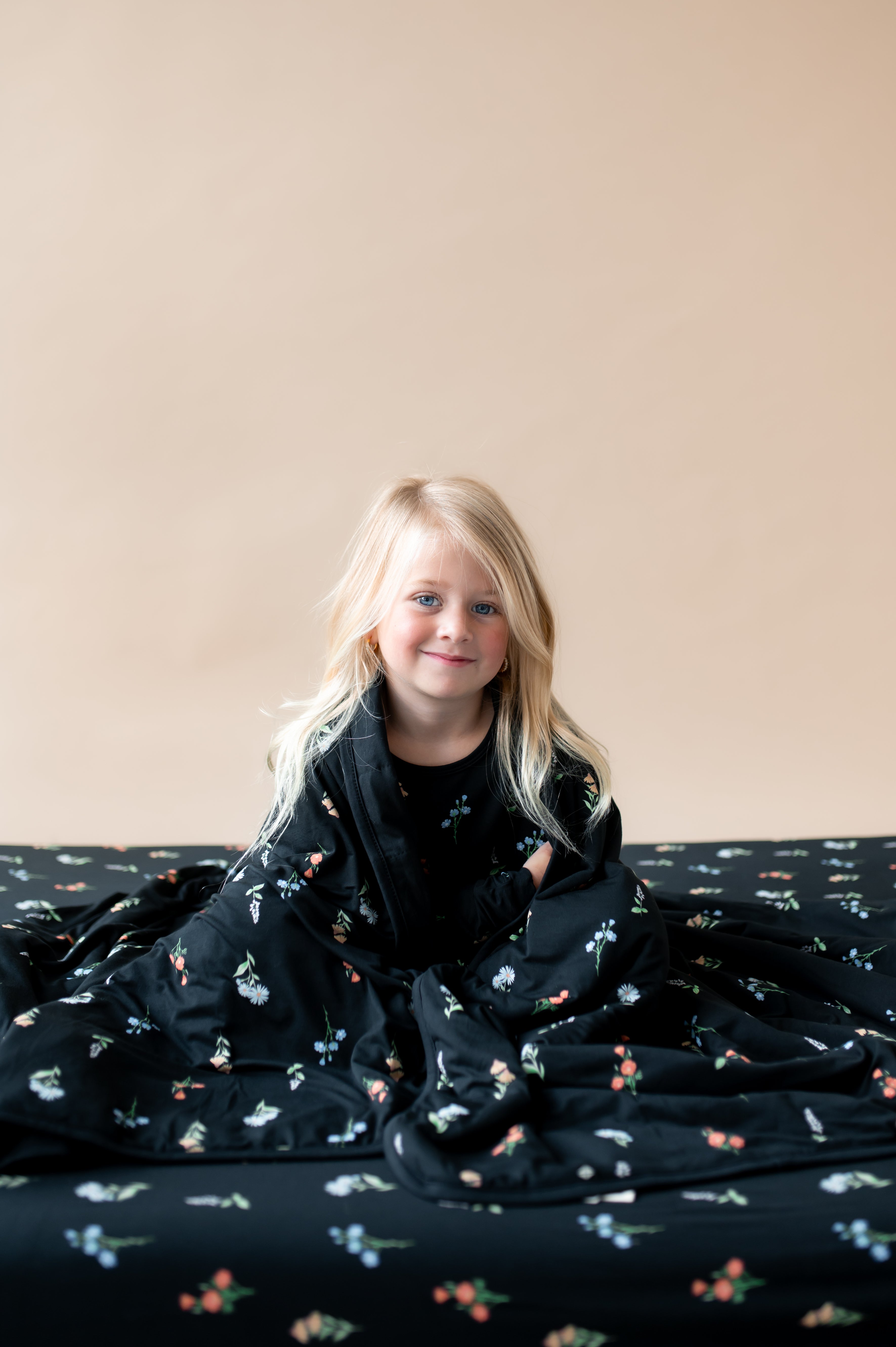 Girl wrapped in Toddler Blanket with Midnight Wildflower 1.0 print sitting on a similar print twin sheet and wearing similar print pajamas on a beige background
