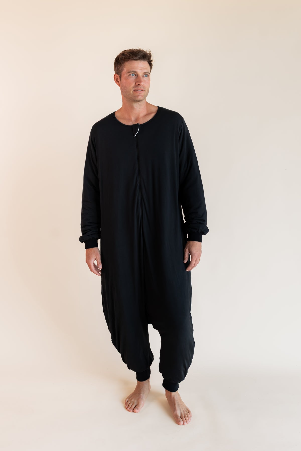 A man wearing a black long-sleeve, one-piece harem-style sleep sack front zipper, standing barefoot against a plain beige studio background.