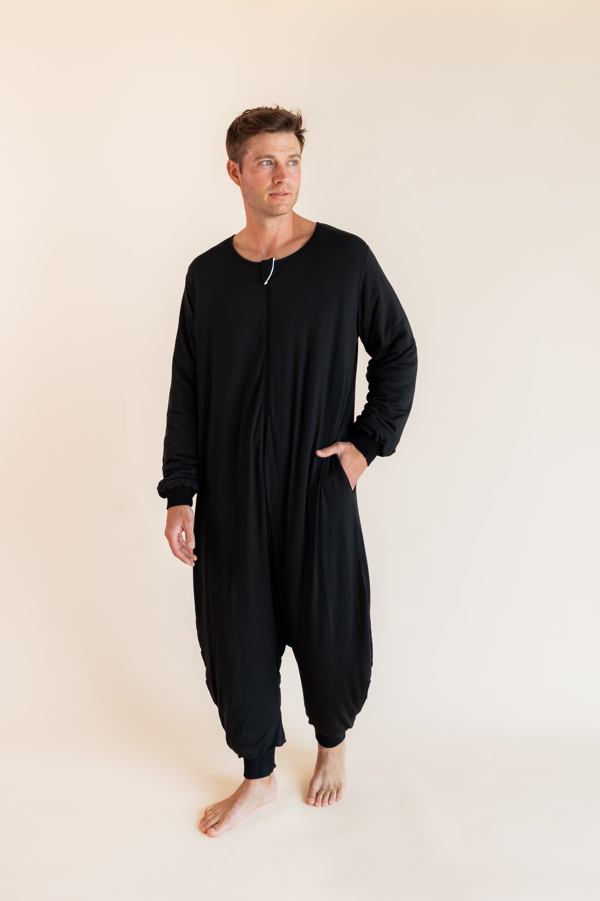 A man modeling a black long-sleeve harem-style sleep sack against a plain beige studio background.