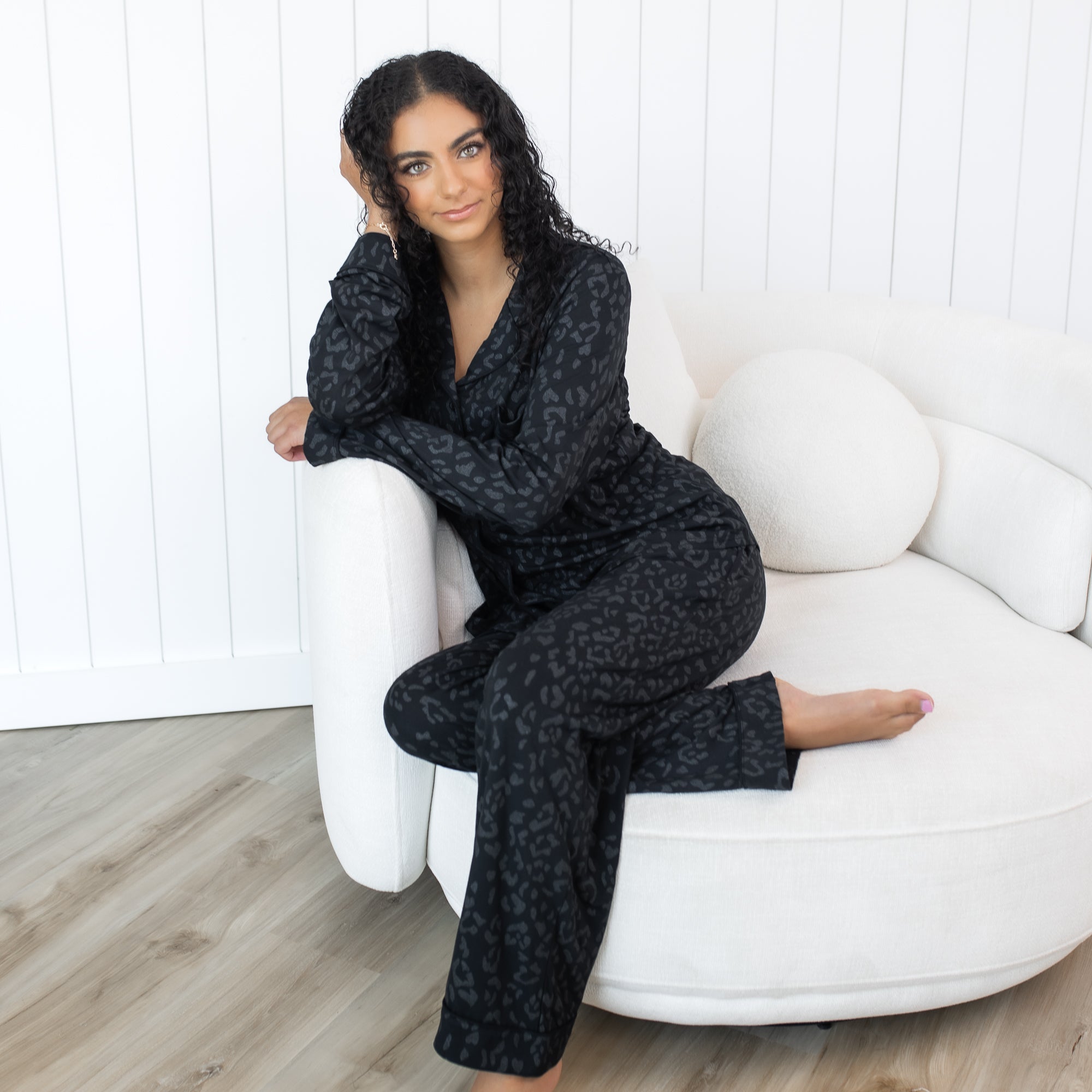 Female sitting on a round sofa wearing the Long-Sleeved Women's Pajama Set in Midnight Leopard in front of a white paneled wall