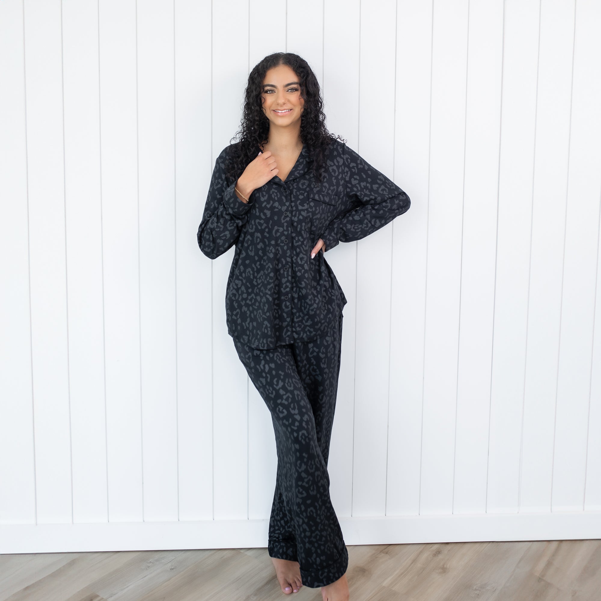 Female standing in front of a white paneled wall wearing the Long-Sleeved Women's Pajama Set in Midnight Leopard