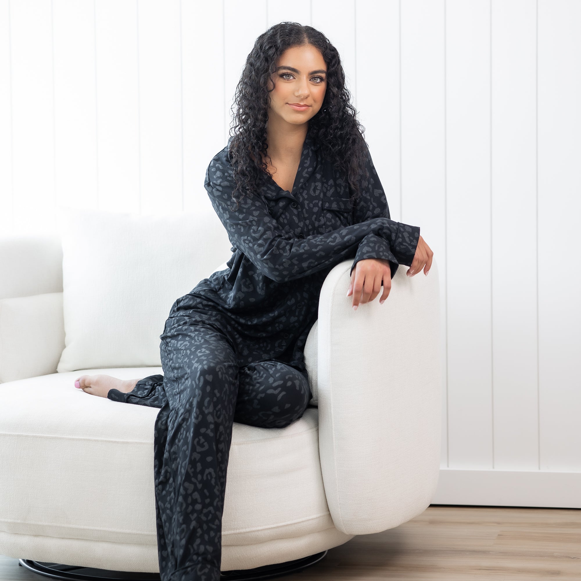 Female sitting on a white sofa with her arms on the sofa arm rest wearing the Long-Sleeved Women's Pajama Set in Midnight Leopard