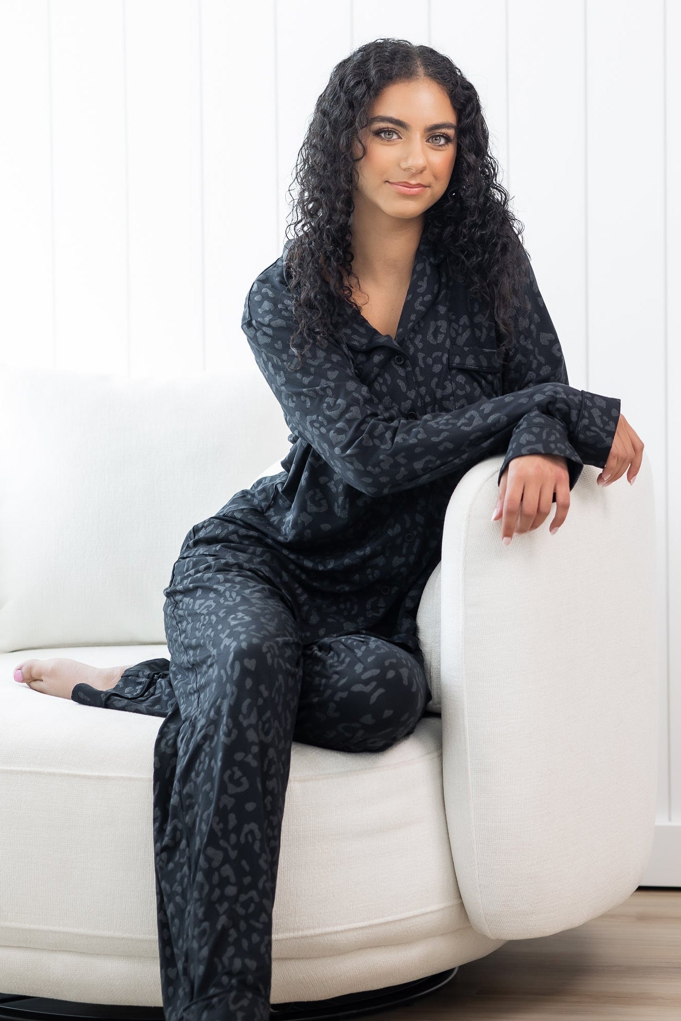 Female sitting on a white sofa with her arms on the sofa arm rest wearing the Long-Sleeved Women's Pajama Set in Midnight Leopard