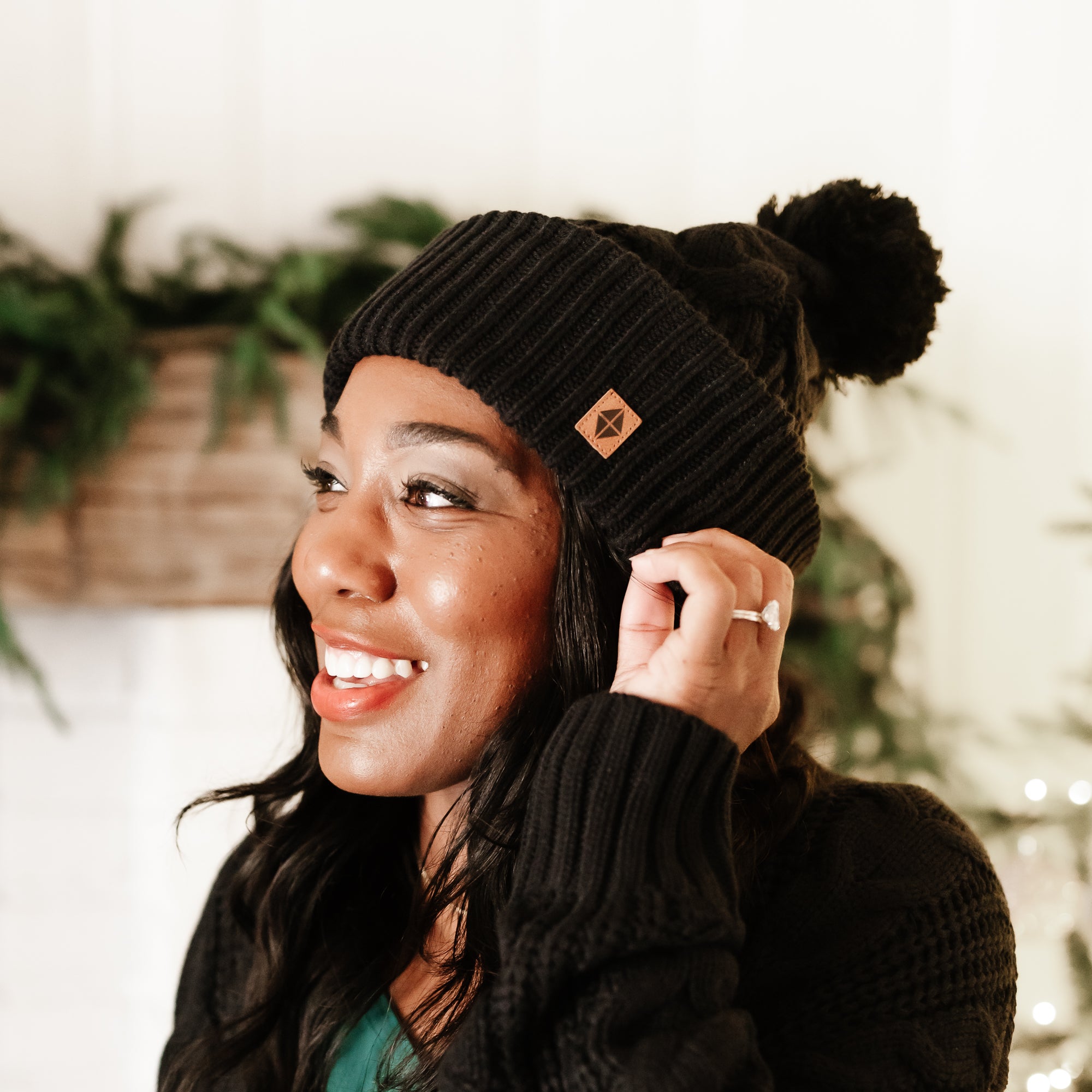 Smiling female wearing the Cable Knit Adult Pom Beanie in Midnight with greenery on a mantel in the background