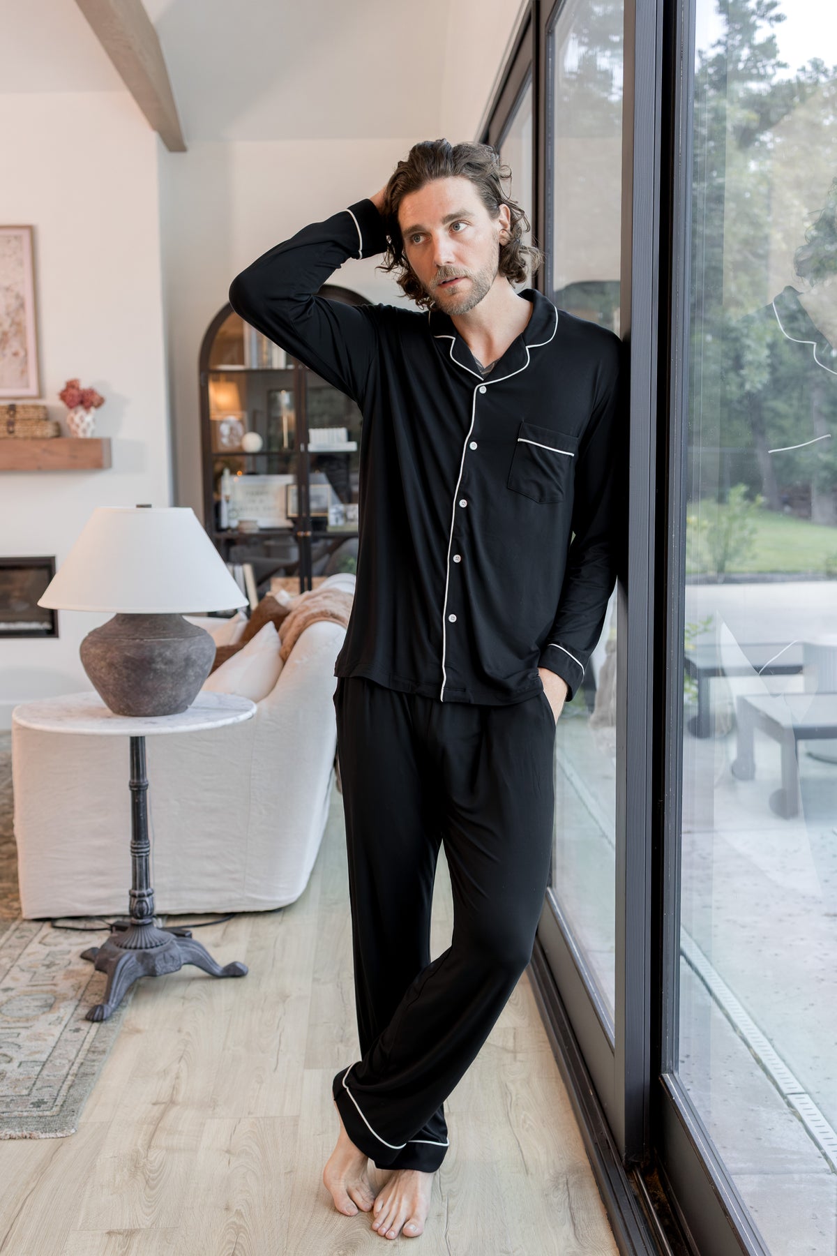 A man wearing a black long-sleeve pajama set with white piping stands barefoot in a bright living room next to large glass doors.