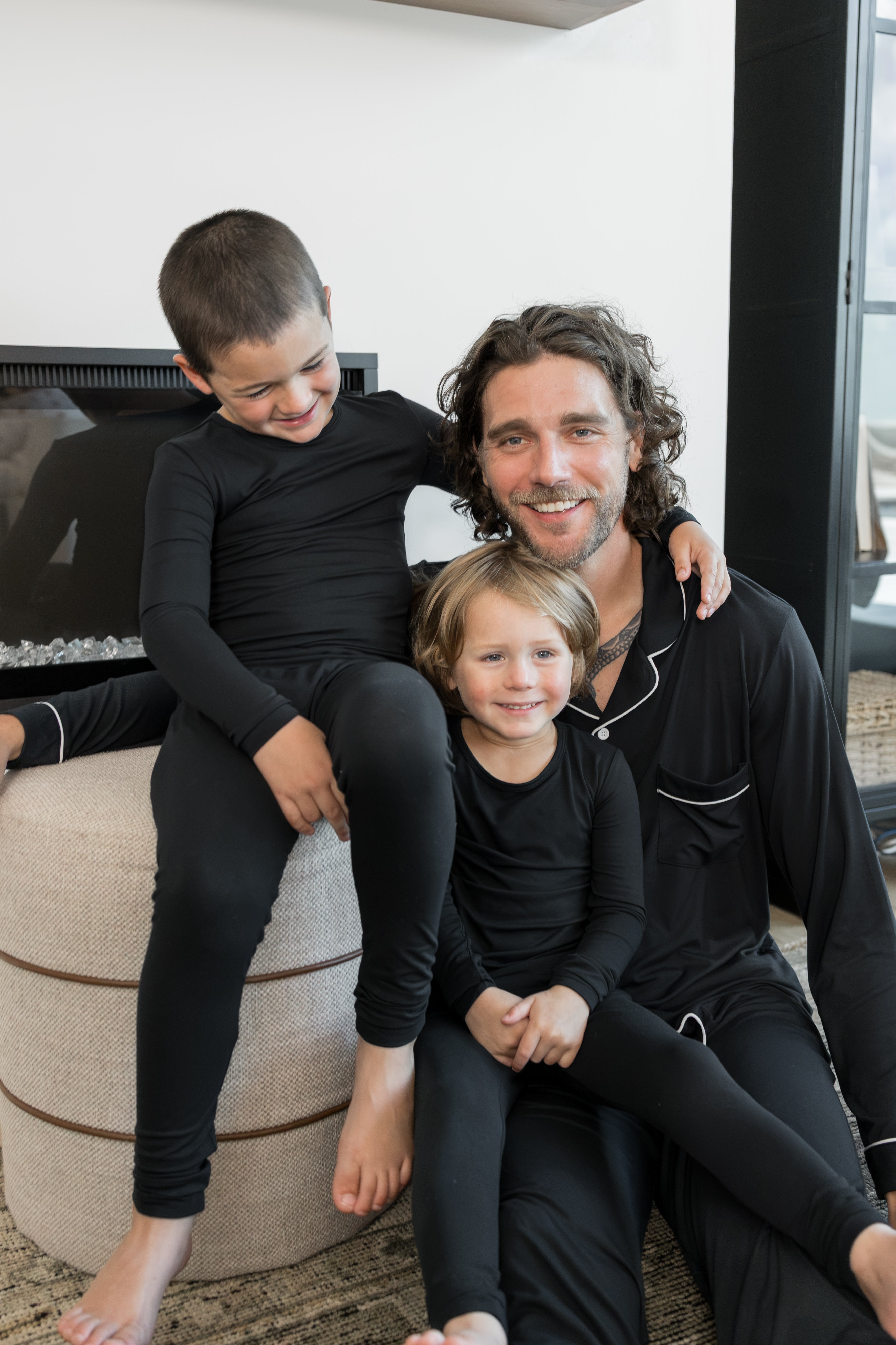 A man and two children wearing matching black long-sleeve pajamas sit together indoors in a cozy living room setting.