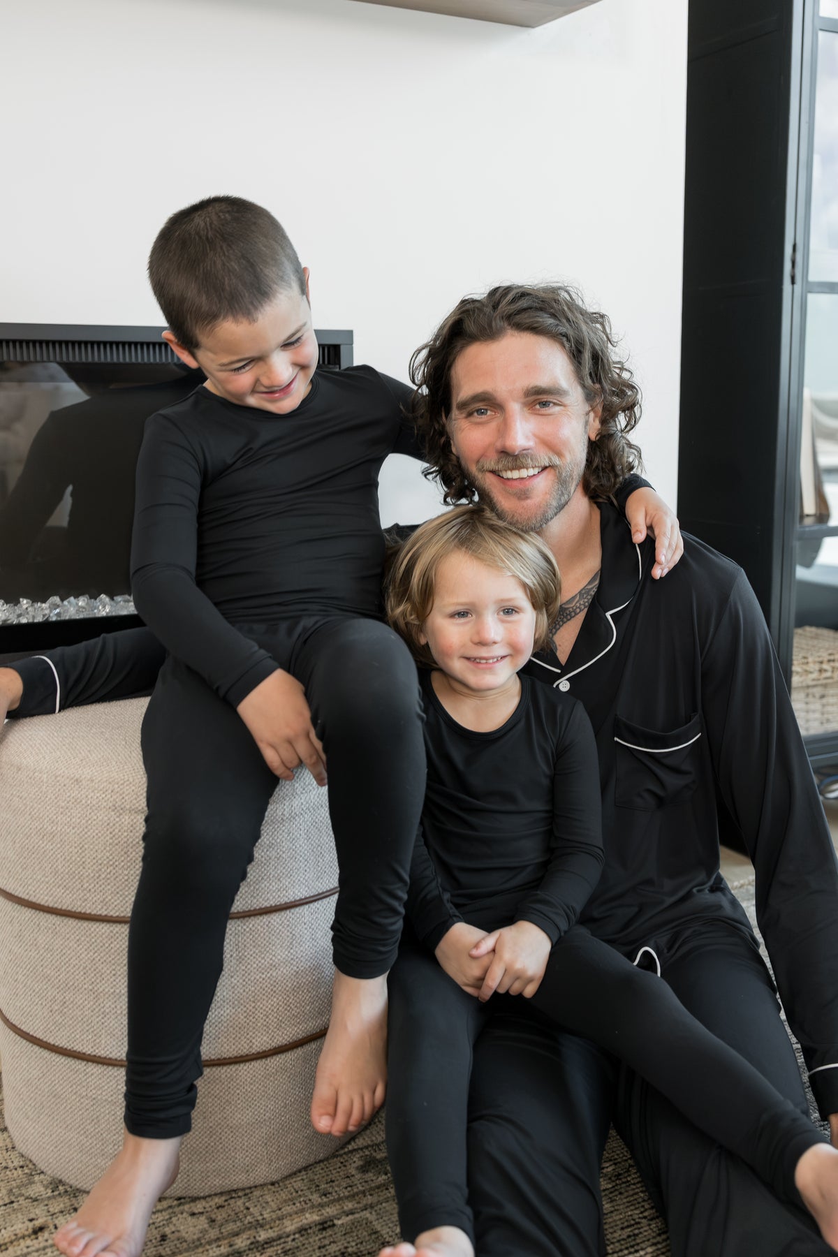 A man and two children wearing matching black long-sleeve pajamas sit together indoors in a cozy living room setting.