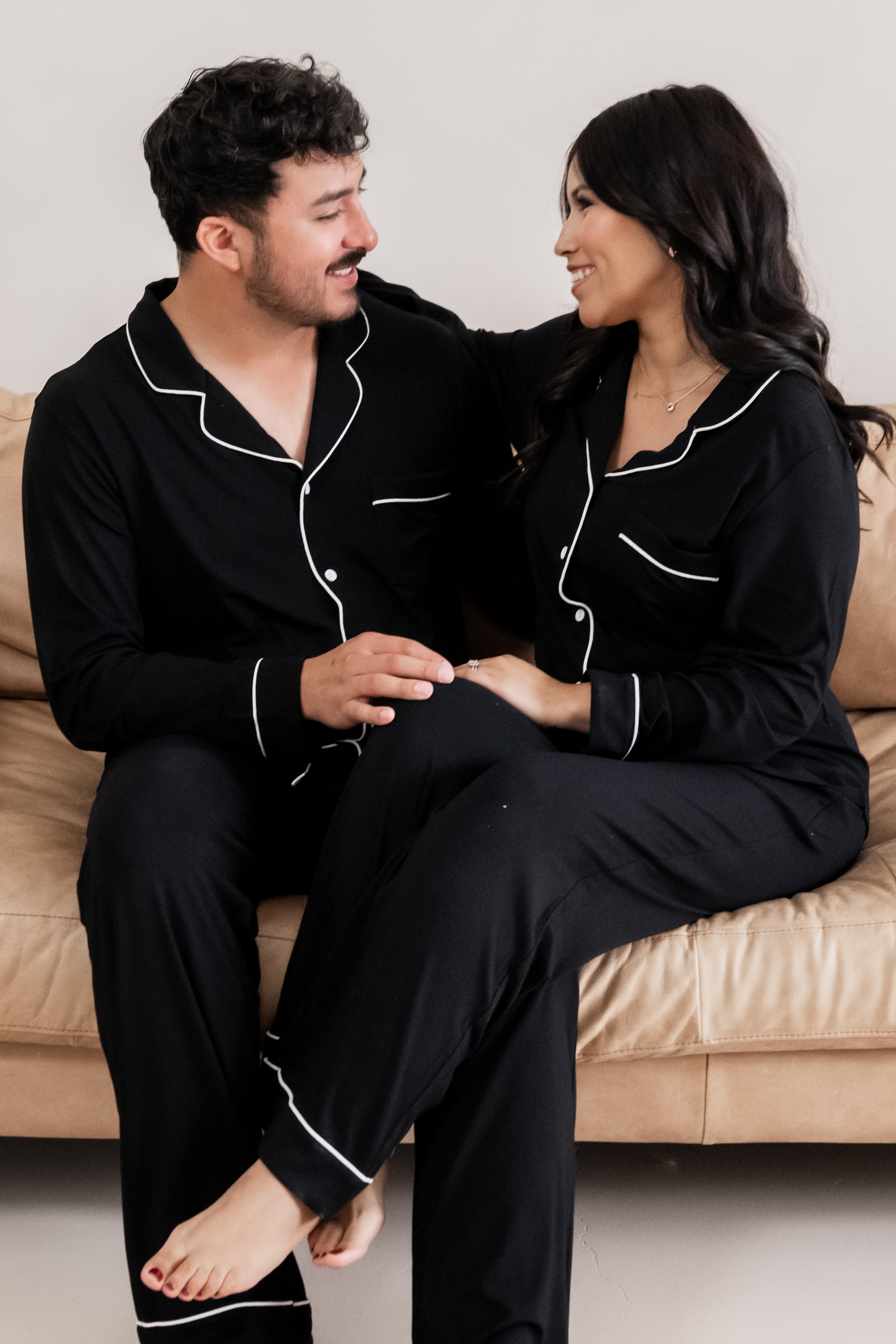 Husband and wife sitting on a couch wearing matching long sleeve midnight pajamas with white trim