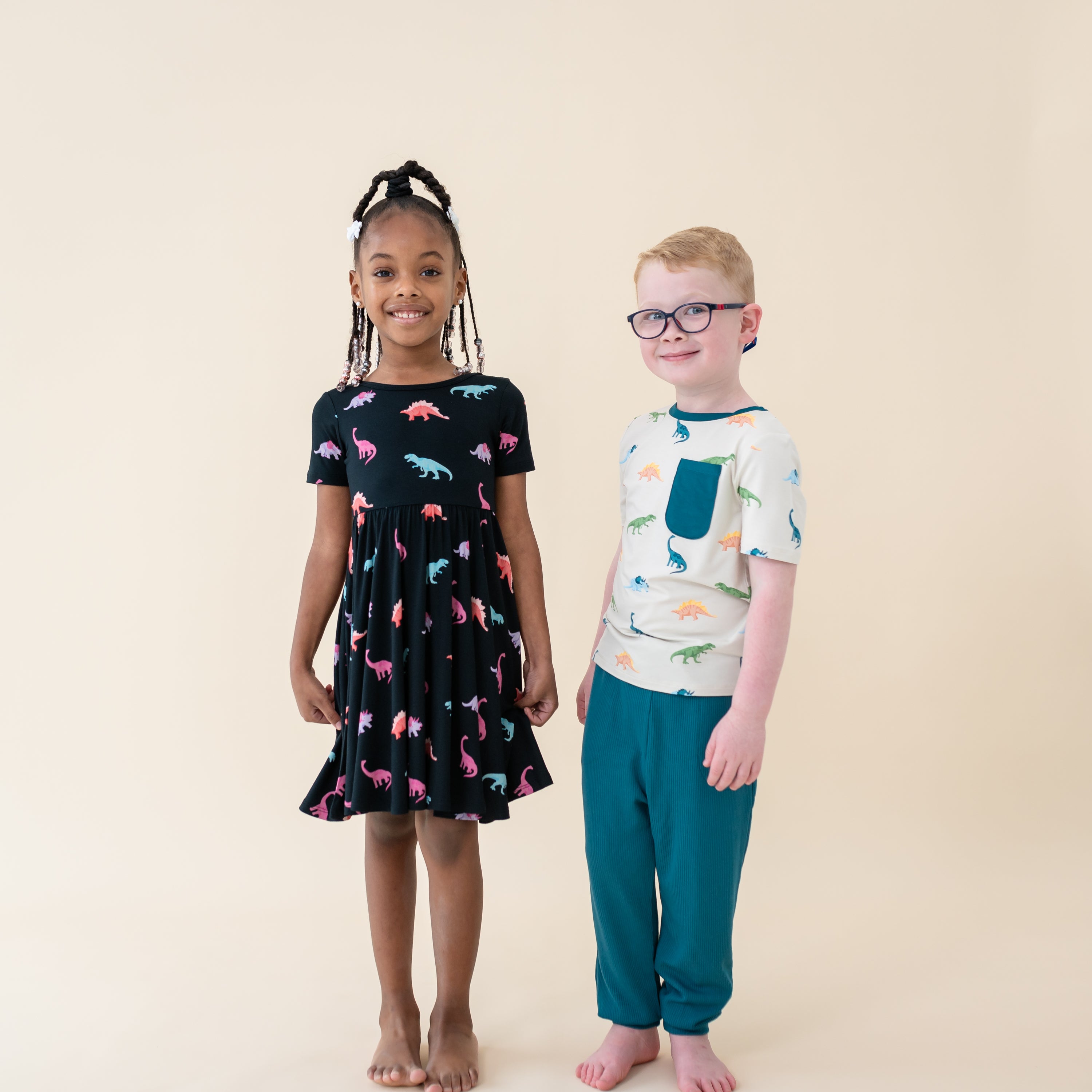 Two smiling children modeling the Roar Collection. Young girl is in Midnight Roar Twirl Dress and young boy is in the Ecru Roar Crew Neck Tee paired with Loch pants