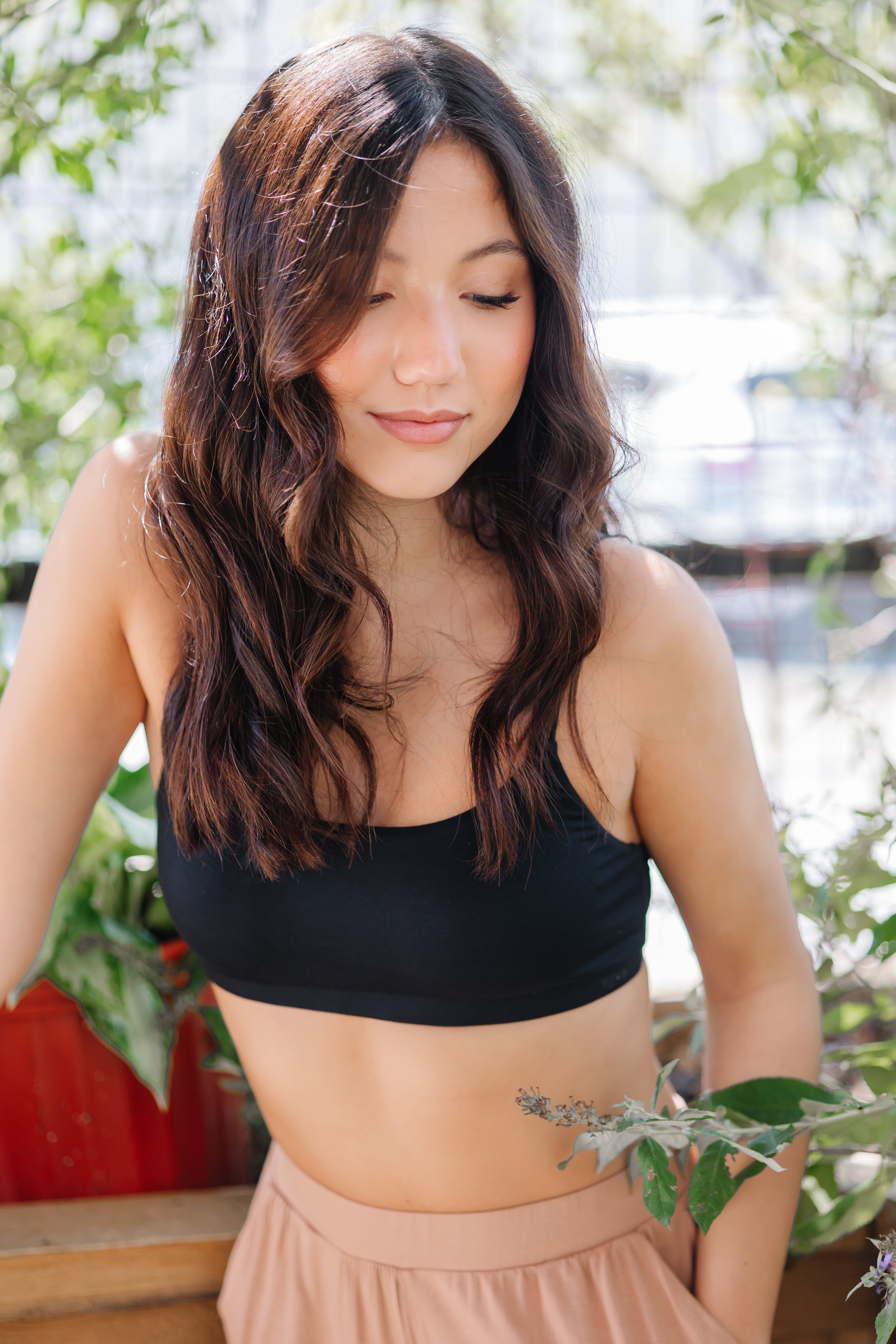 Woman wearing a black bralette and beige pant with plants in the background