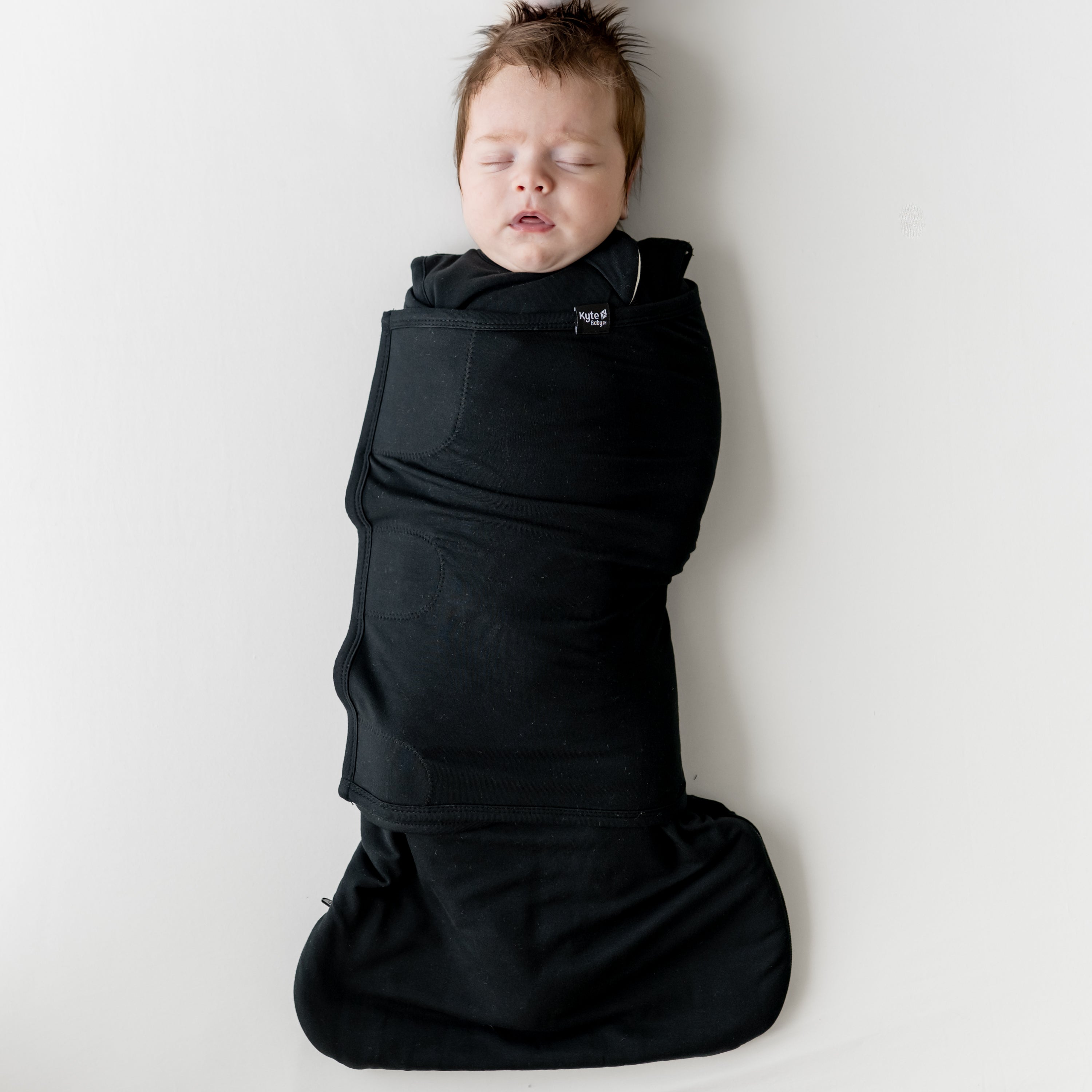 Sleeping infant laying on a light neutral background swaddled in the Sleep Bag Swaddler in Midnight 1.0