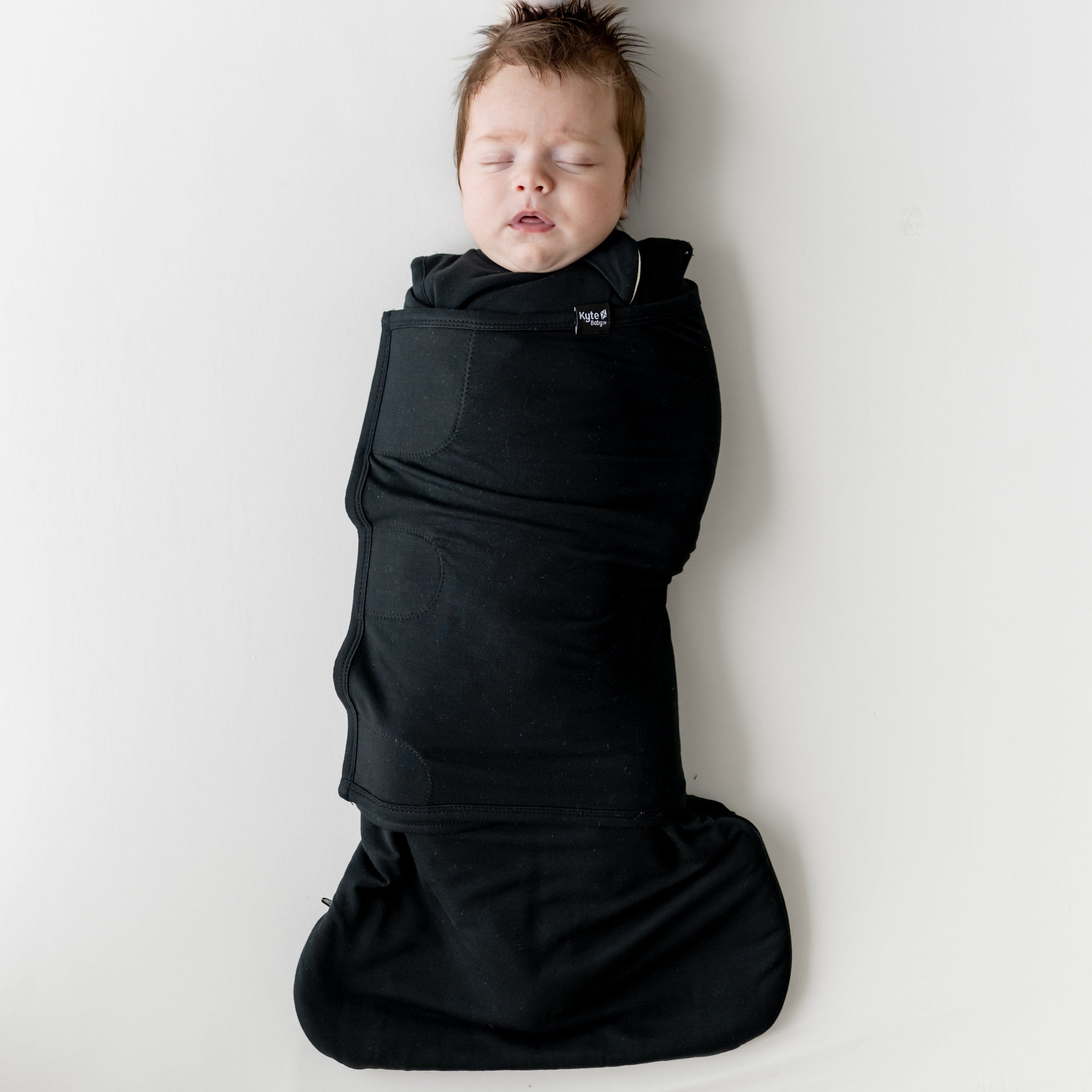 Sleeping infant laying on a light neutral background swaddled in the Sleep Bag Swaddler in Midnight 1.0