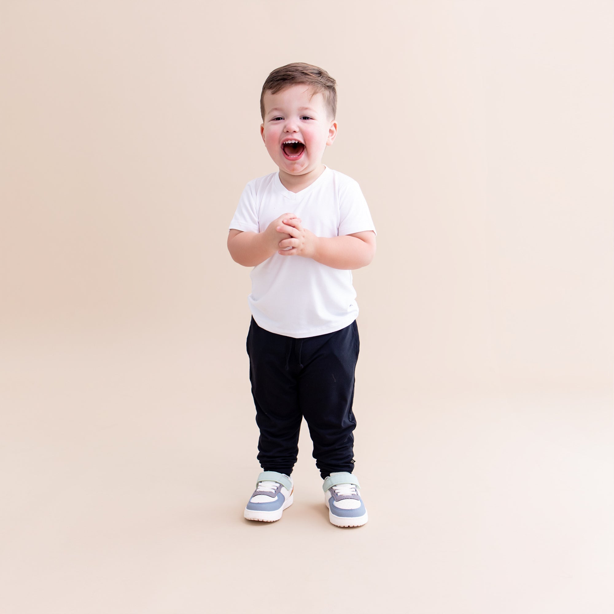 Smiling toddler standing in front of a light beige background wearing the Toddler Jogger Pant in Midnight with a white v-neck shirt