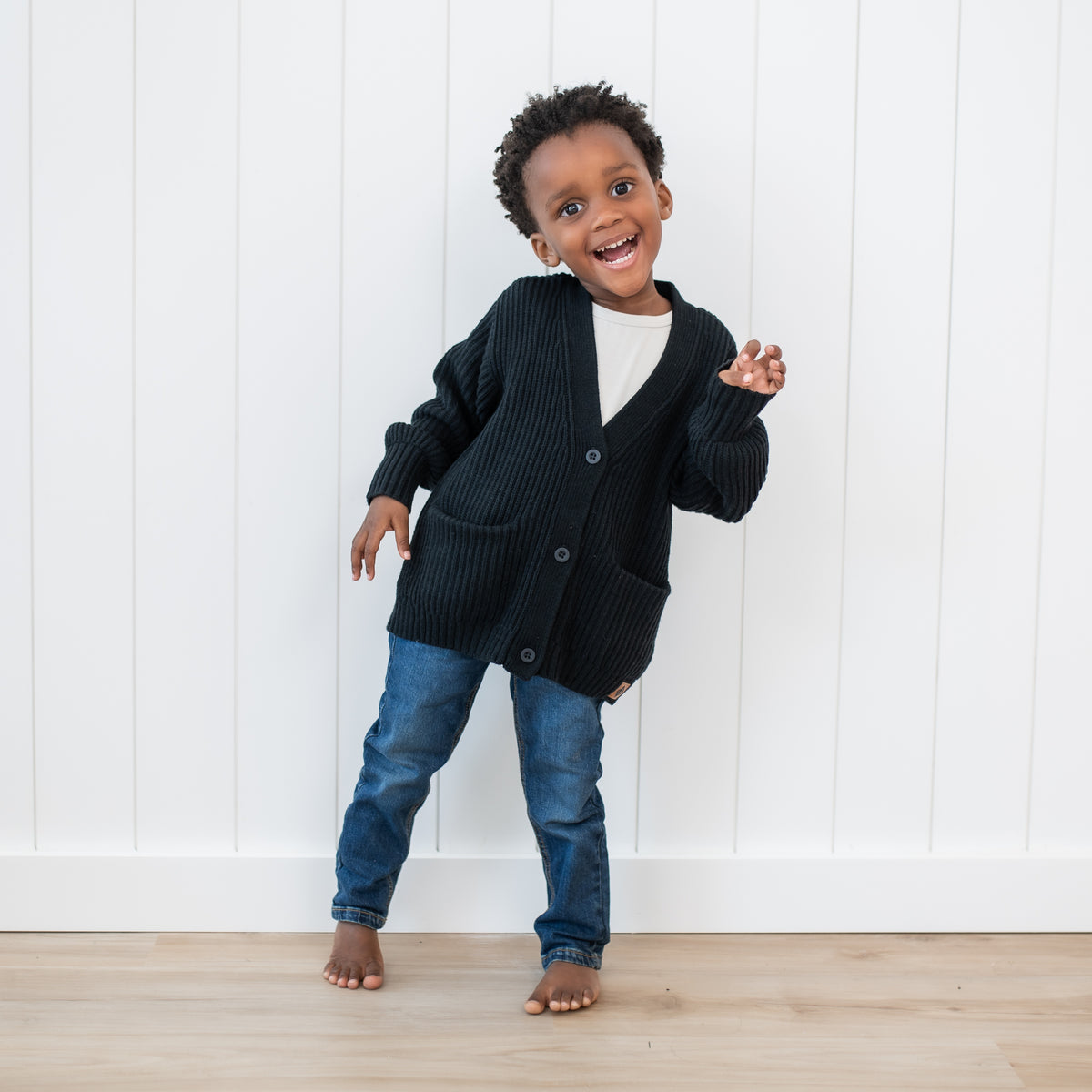 Toddler wearing the Chunky Knit Oversized Cardigan in Midnight with medium wash jeans in front of a white paneled wall