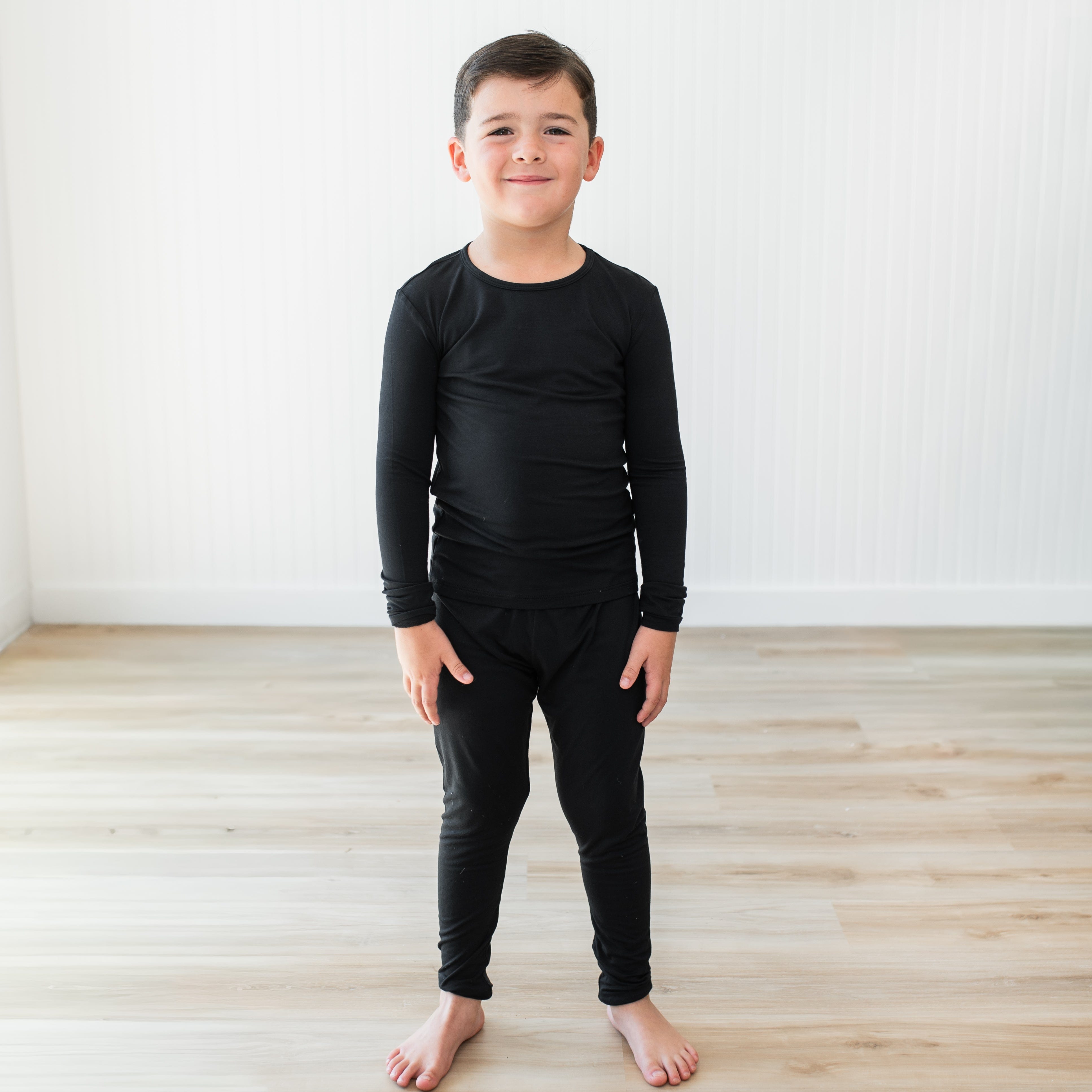 Child wearing a black long-sleeve shirt and pants pajama set standing on a wooden floor with a white wall background