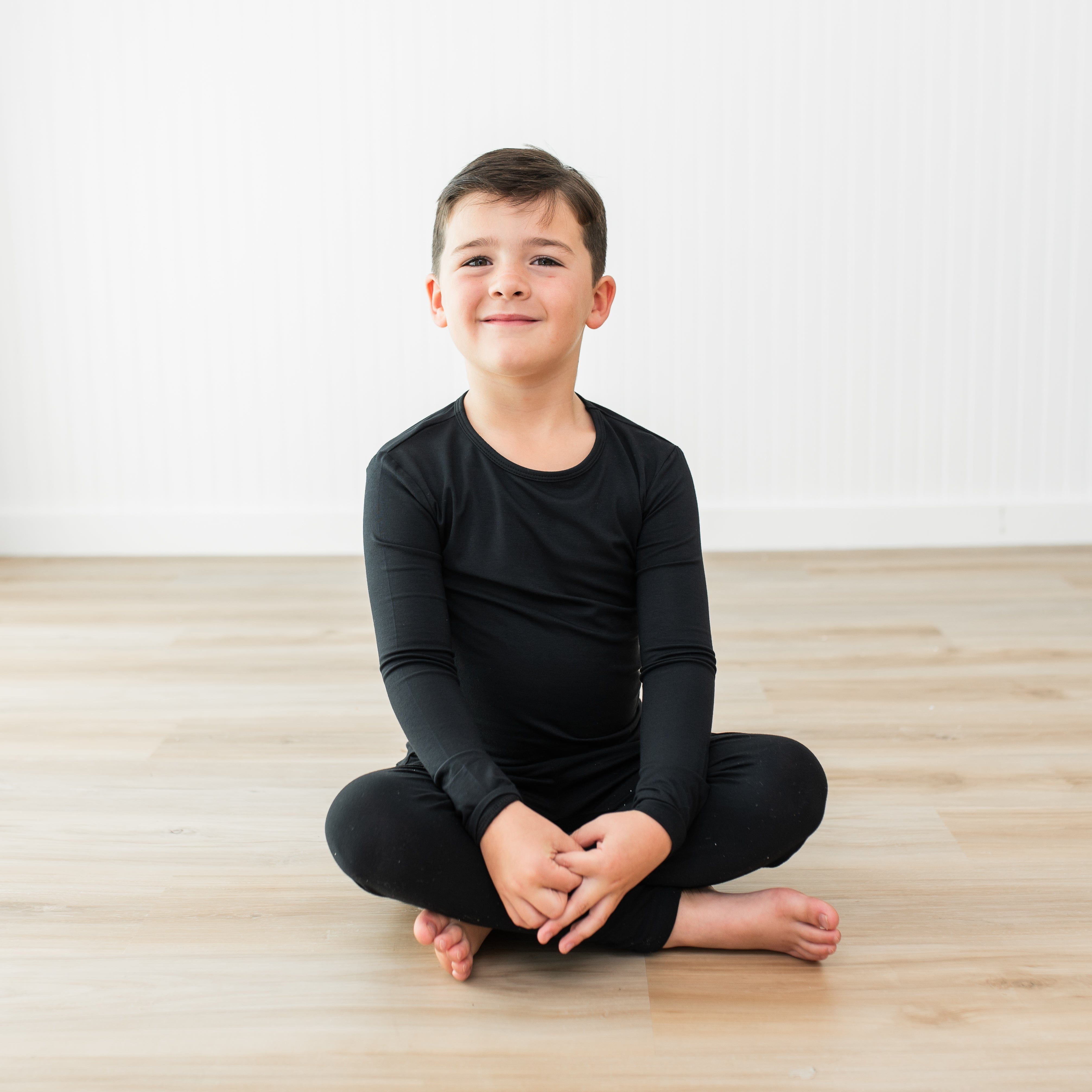 Child sitting cross-legged on a wooden floor wearing a black outfit.