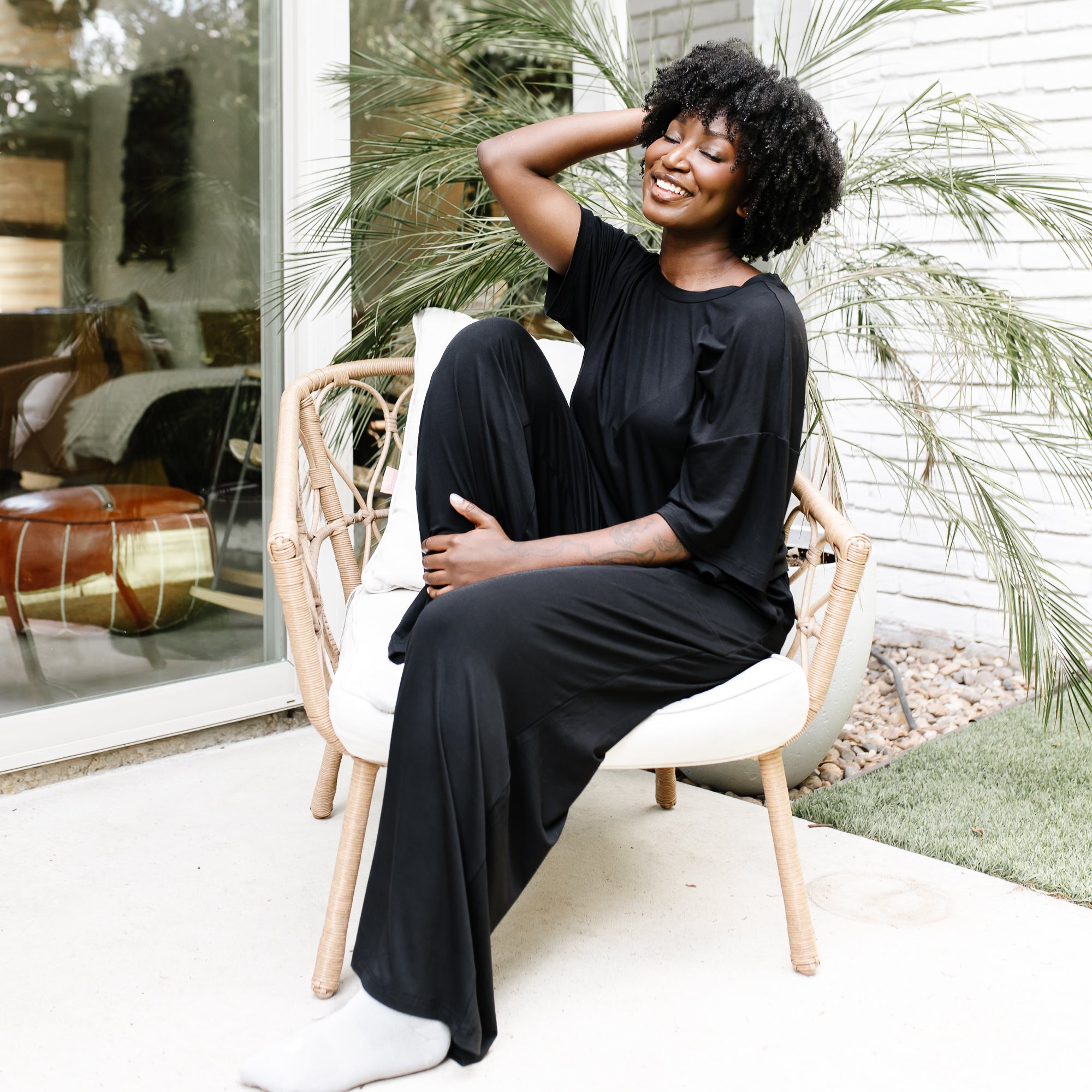 Smiling female sitting on a wicker chair on white cushion with her eyes closed wearing the Women's Wide Leg Pant in Midnight with matching women's short sleeve tee