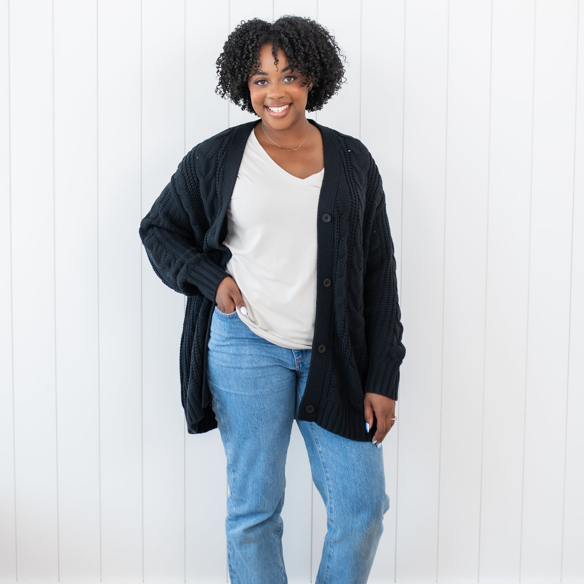 Smiling female standing with one hand in her jean pocket wearing the Cable Knit Adult Cardigan in Midnight