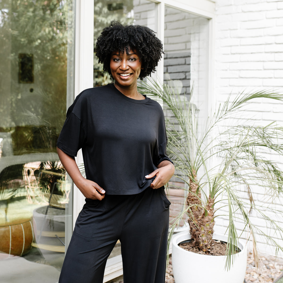 Smiling female model standing in front of a white brick house wearing the Women's Short Sleeve Tee in Midnight with matching wide leg pants