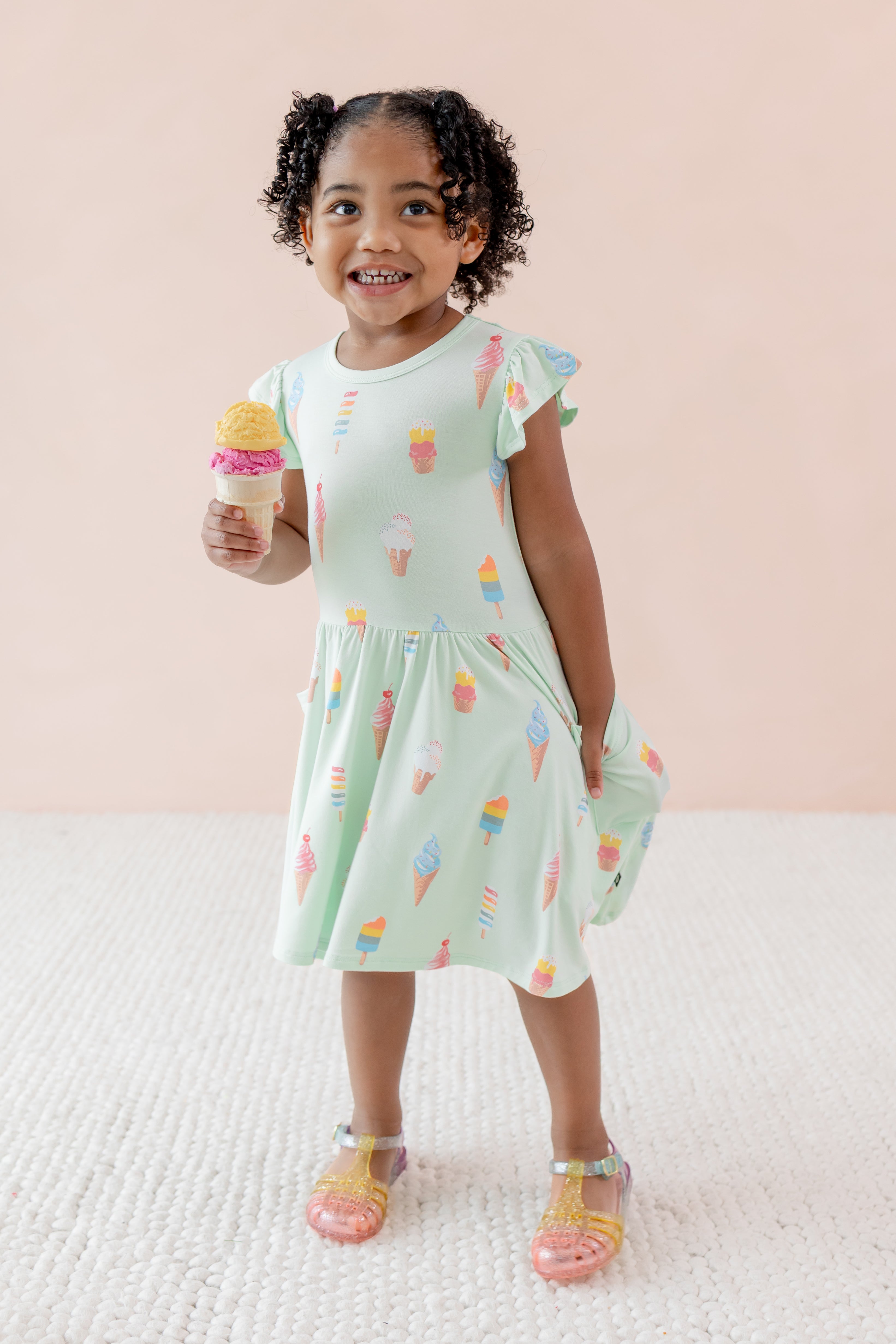 Child posing in Pocket Dress with Mint Ice Cream print and sandles on a beige background holding an ice cream